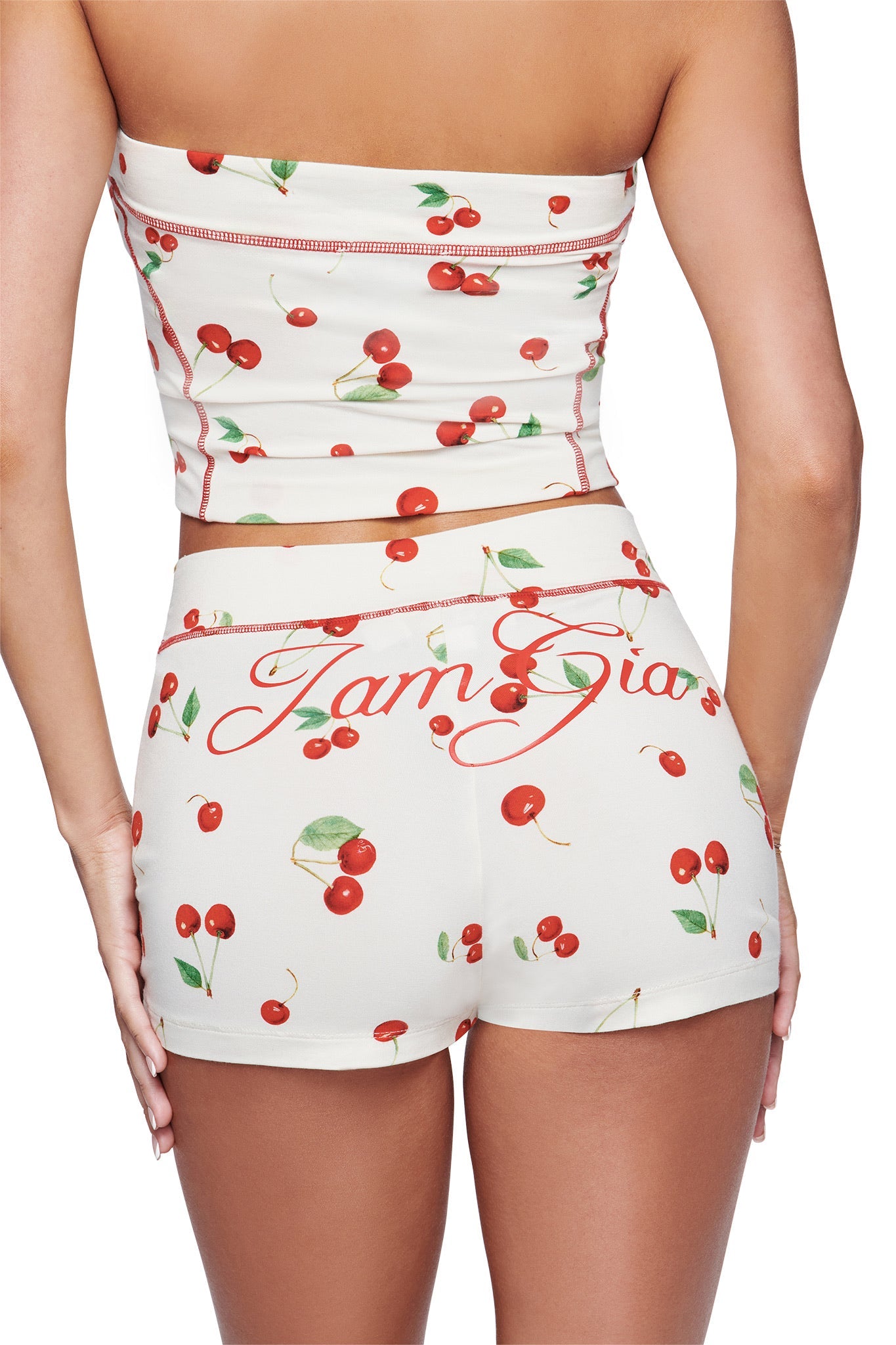A woman is shown from the back wearing a matching cream-colored cherry print tube top and the I.AM.GIA BLARE SHORT - CHERRY ON TOP mini shorts. The shorts feature an elastic waistband with “Jam Gia” written in red script across the back, offering a stylish nod to I.AM.GIA’s signature look.