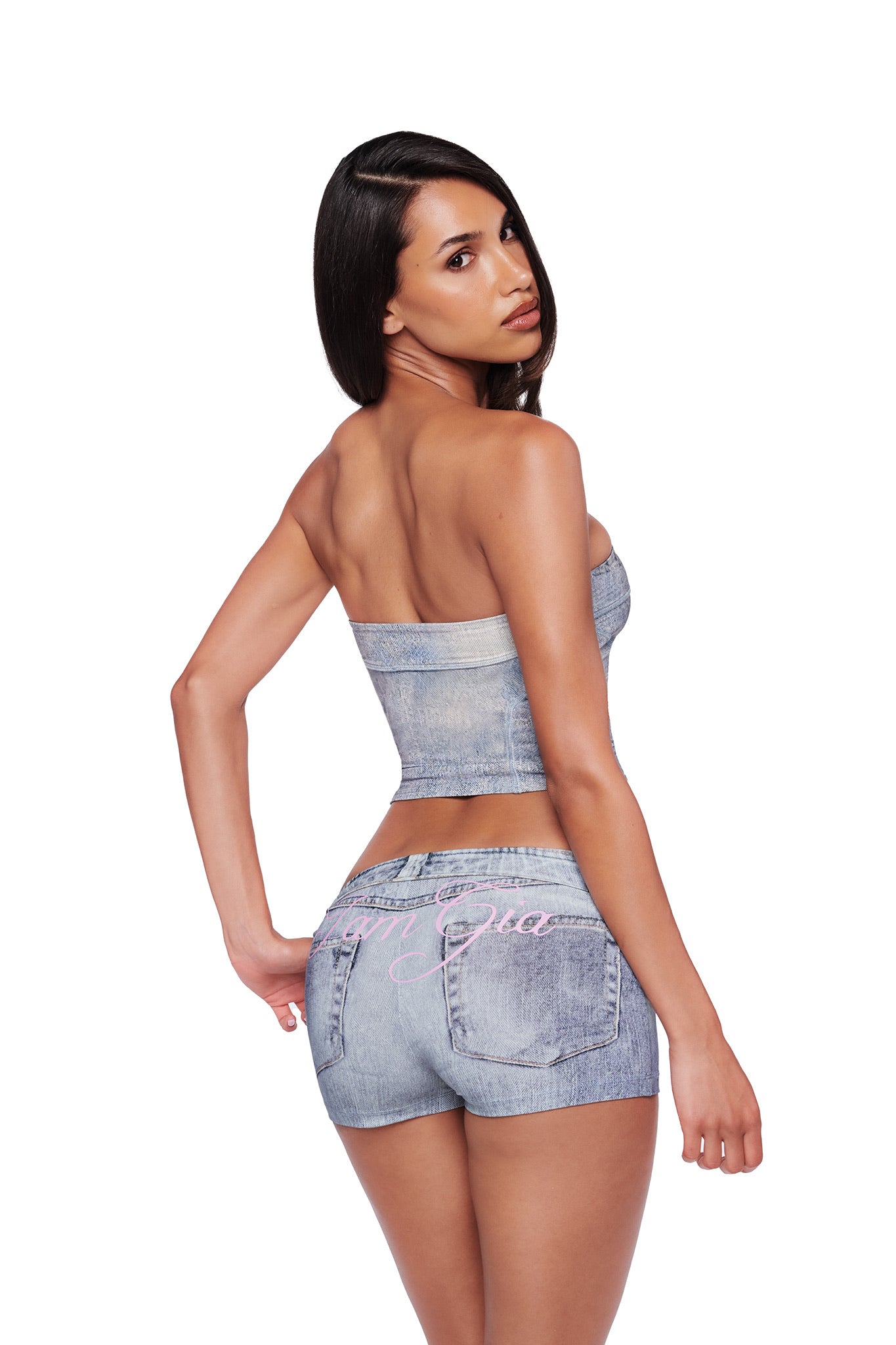 A woman with long dark hair poses in a strapless denim top and the BLARE SHORT - DENIM TROMPE L’OEIL by I.AM.GIA, standing against a white background and glancing over her shoulder at the camera.
