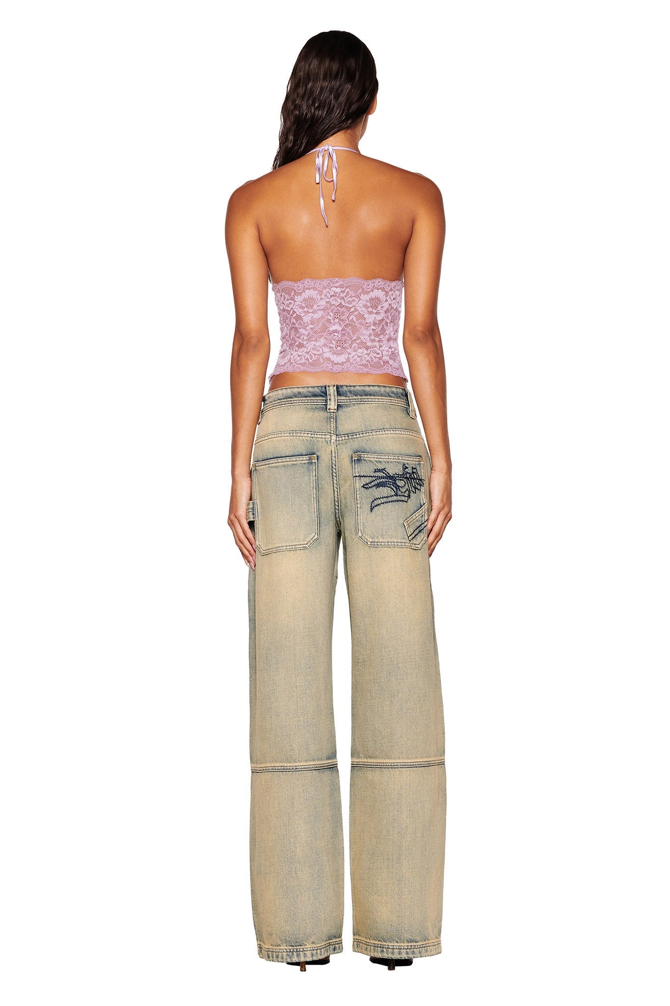 A person with long hair stands facing away, wearing the GEM HALTER TOP in lilac purple by I.AM.GIA and baggy light-colored jeans featuring visible stitching and a blue embroidered design on the back pocket, set against a plain white background.