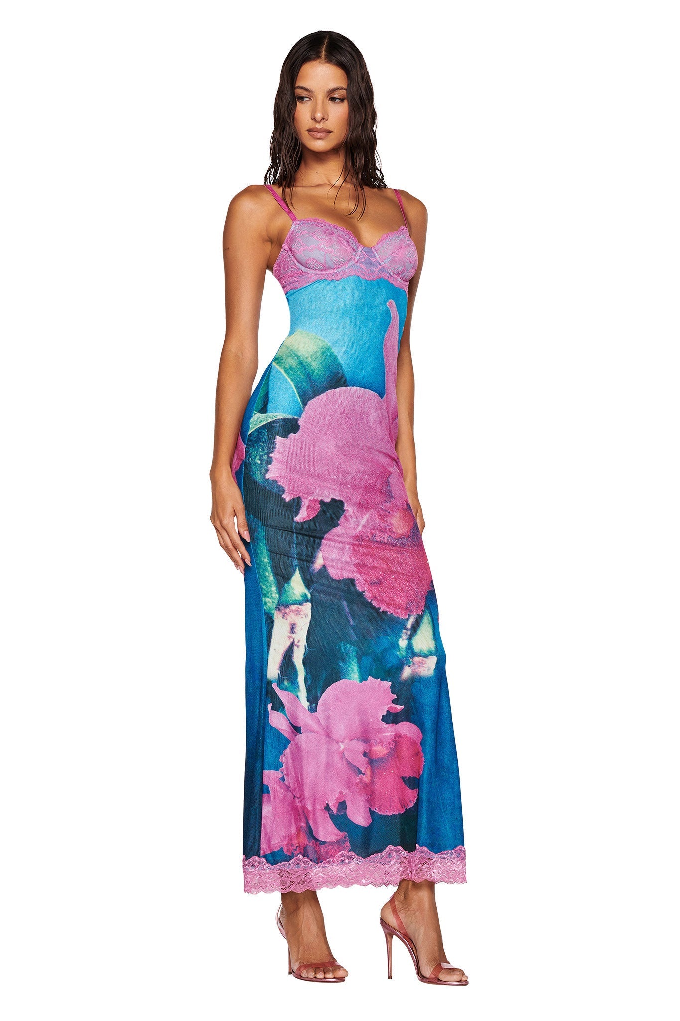 A woman with long hair is showcasing the I.AM.GIA Calypso Maxi Dress in a blue orchid floral print. She stands with one hand to her side, complementing the maxi-length dress's fitted bodice and lace hem details, paired elegantly with high-heeled shoes.