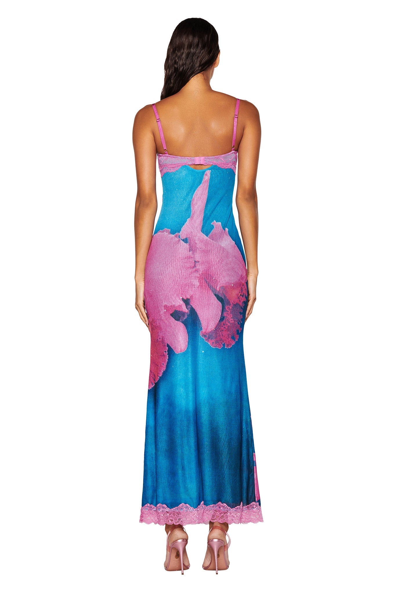 A woman is wearing the I.AM.GIA Calypso Maxi Dress, which showcases a blue background adorned with a large pink orchid print. The dress is sleeveless and maxi-length, featuring lace trim at the hem. She pairs it with pink high-heeled sandals and stands with her back to the camera.