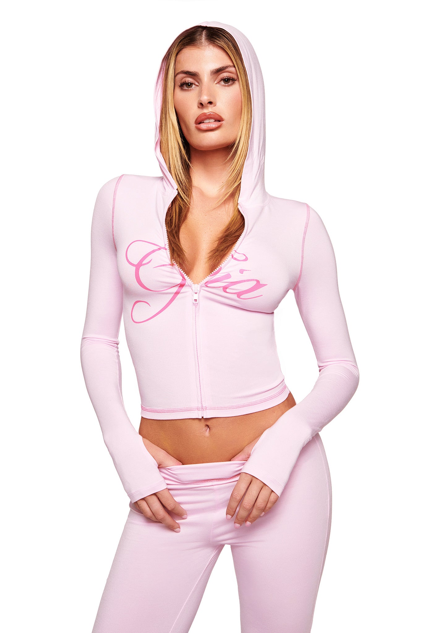 A woman poses against a white background in the I.AM.GIA BLARE HOODIE - PINK, a pastel pink bodycon cropped hoodie with "Gia" on the front, paired with matching pink pants, hood up and hands near her waist.