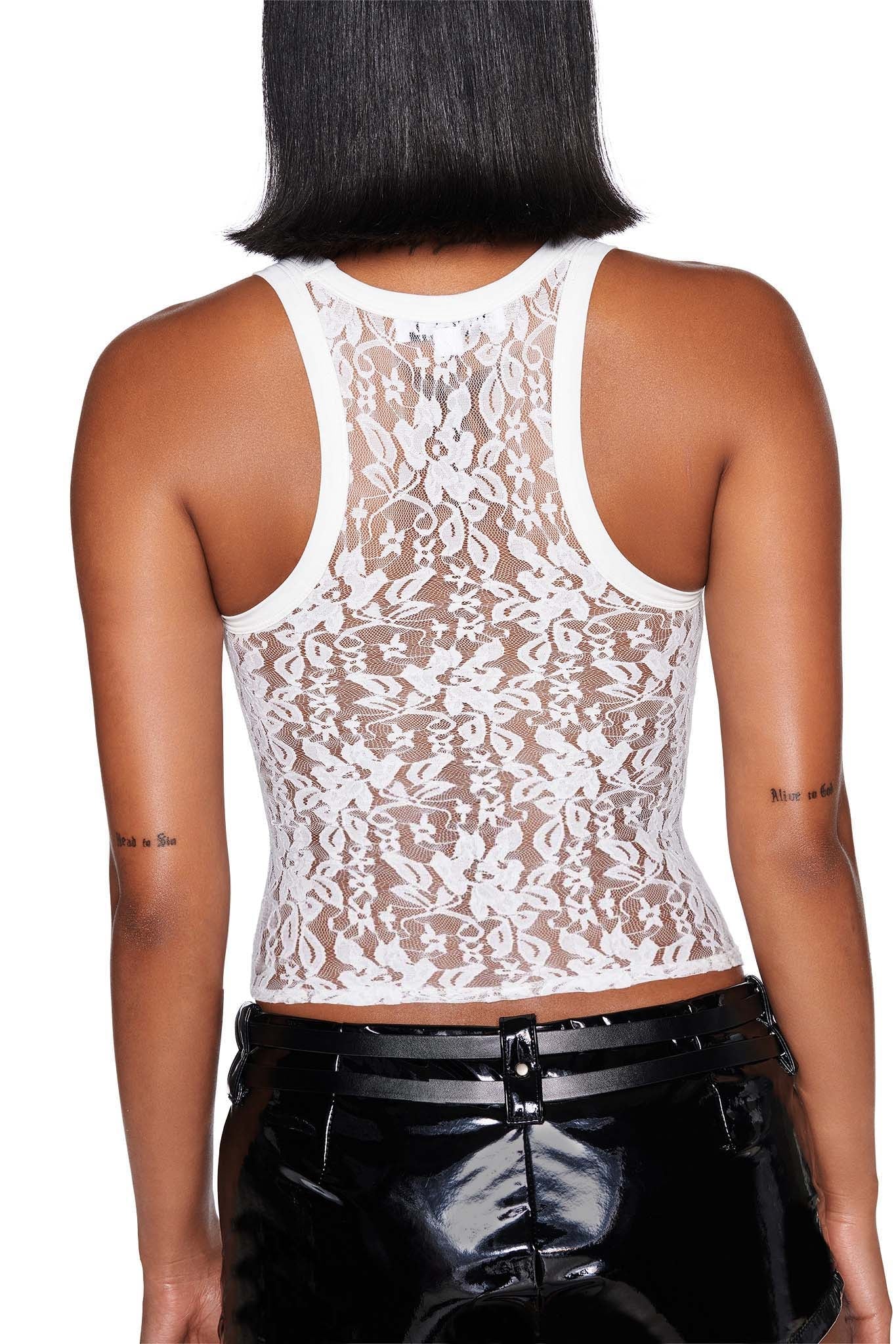 A person wearing the SAGITTARIUS ZODIAC TANK - WHITE from I.AM.GIA, a lace racer-style top with intricate patterns that reveals their back. They have long hair and are paired with shiny black bodycon pants, displaying tattoos on both arms.