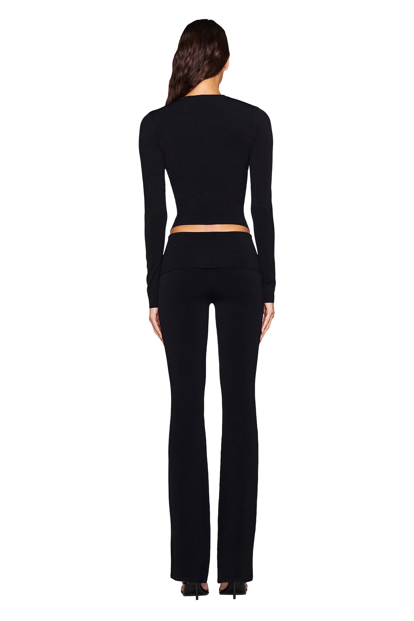 A long-haired person in a fitted black top and I.AM.GIA's AUDRINA PANT 2.0 - BLACK, featuring a flared leg and foldover waistband, stands back to the camera on sleek black high heels against a plain white backdrop.