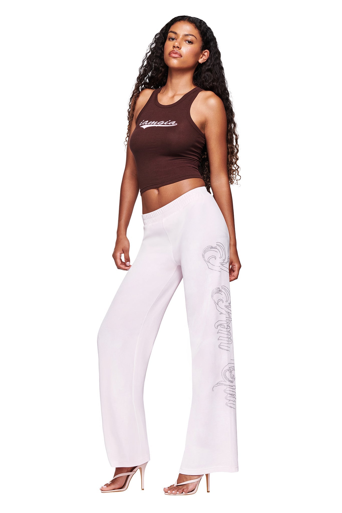 A woman with long curly hair wears the I.AM.GIA PENELOPE TANK in brown and light pink embroidered wide-leg pants, paired with clear high-heeled sandals, standing against a white background.