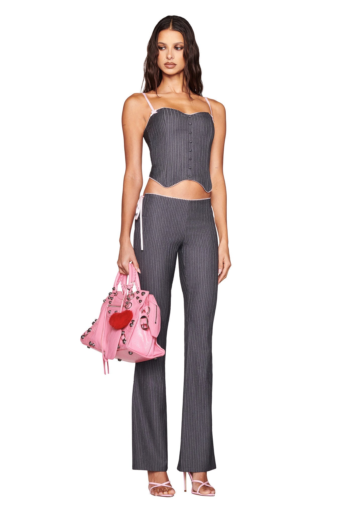 A woman in the FLORIAN CORSET - GREY by I.AM.GIA, paired with matching pants and a pink floral handbag featuring a red heart, wears pink high-heeled sandals. She has long wavy hair against a plain white background.