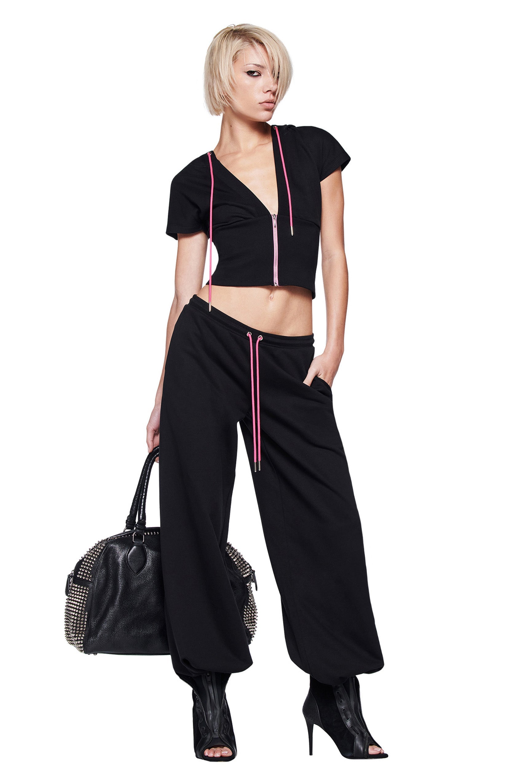 A model with short blonde hair showcases the I.AM.GIA ARLO TRACK PANT in black, styled with a cropped top featuring a pink zipper, black peep-toe heels, and a studded handbag, set against a white background.