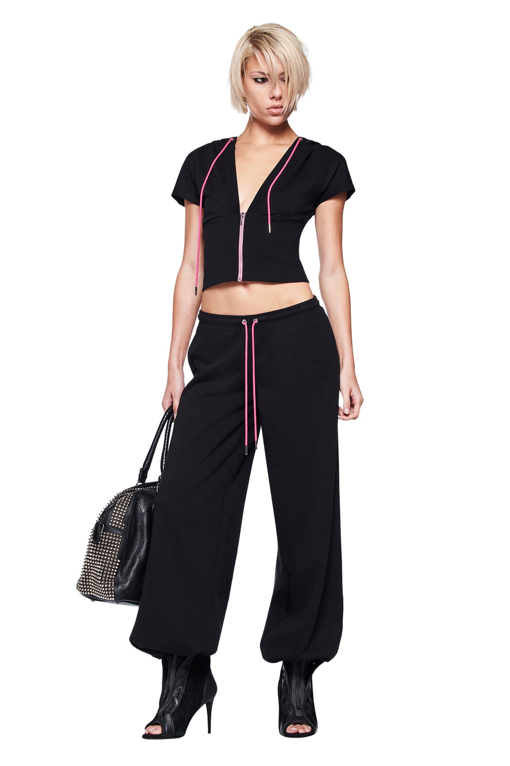 A person with short blonde hair wears the I.AM.GIA ARLO TRACK JACKET - BLACK as a black v-neck zip-up crop top with pink trim, paired with loose black pants with pink drawstrings, open-toe heels, and a studded handbag against a white background.