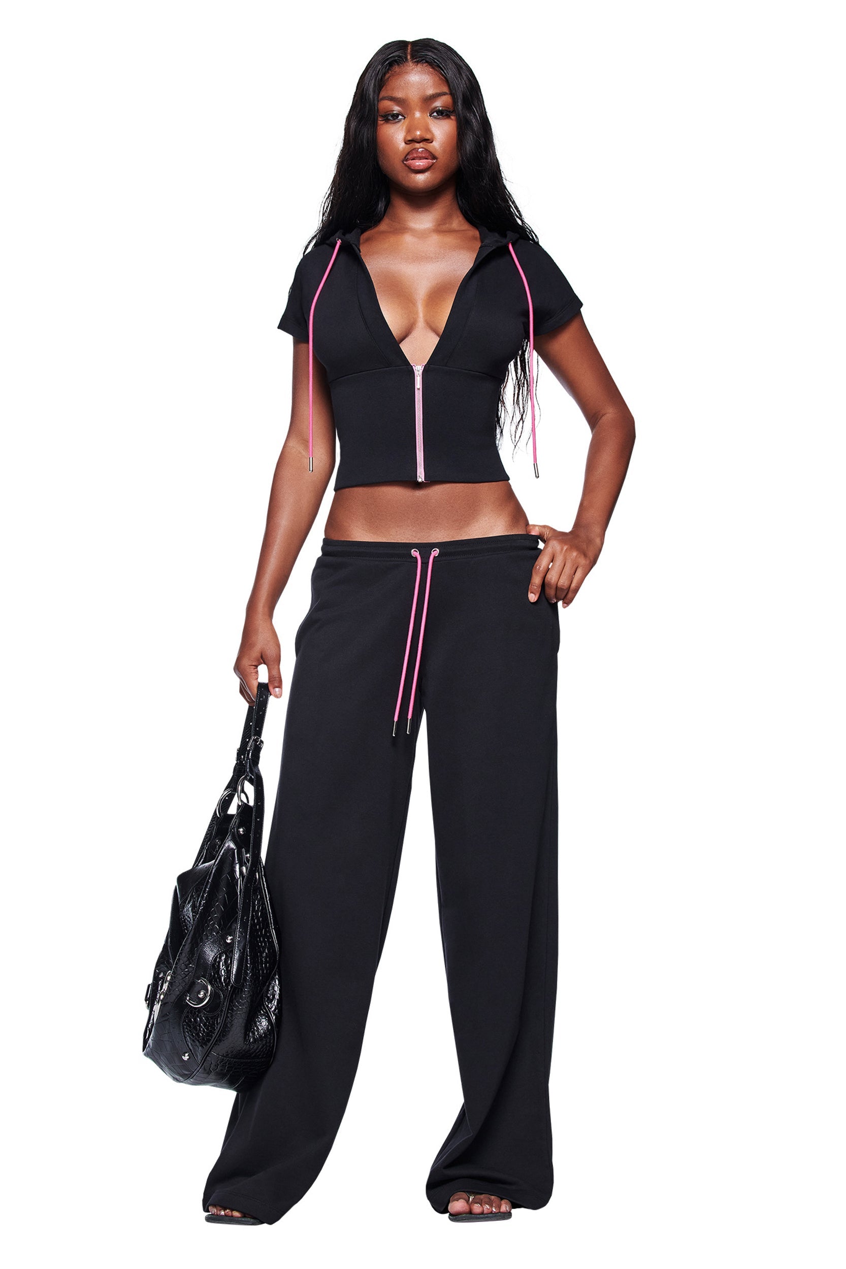 A woman stands confidently in I.AM.GIA’s ARLO TRACK PANT in black, paired with a matching zip-up top. She holds a large black handbag, her long wavy hair flowing, all set against a clean white background.
