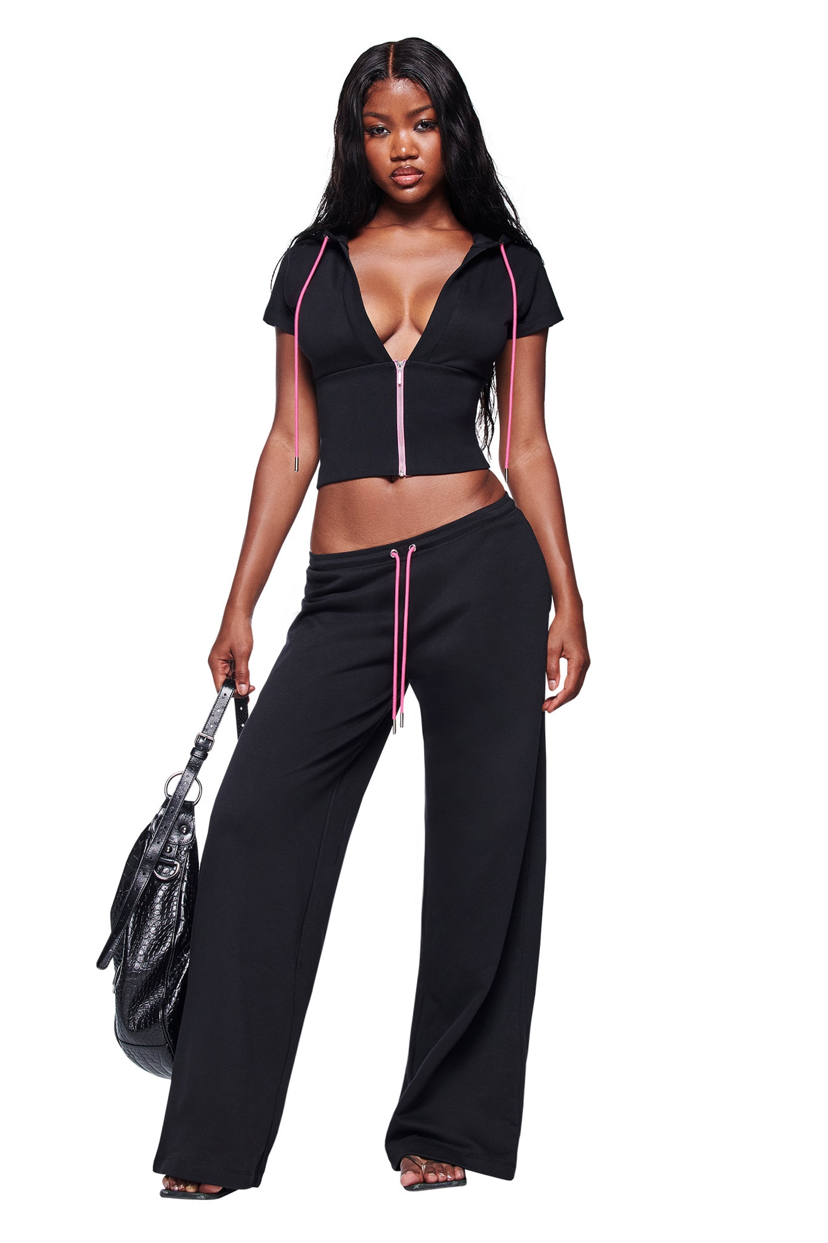 A woman with long, wavy hair stands barefoot against a white background, wearing the I.AM.GIA ARLO TRACK PANT - BLACK with pink drawstrings and a black, short-sleeved zip-up crop top, holding a large black handbag.