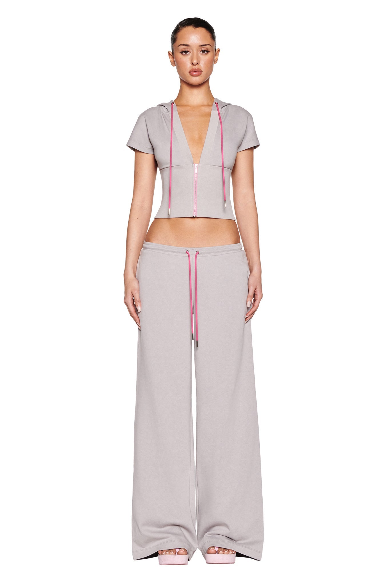 Someone is wearing a light gray, short-sleeved cropped hoodie paired with I.AM.GIA's ARLO TRACK PANT - GREY : PINK wide-leg design, featuring pink drawstrings. The stylish low-rise ensemble stands out against the white background.