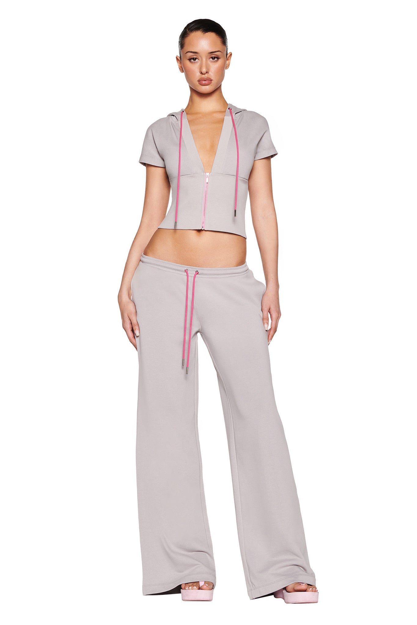 A person wearing the I.AM.GIA ARLO TRACK PANT - GREY : PINK, featuring a stylish light gray zip-up short-sleeved top and matching low-rise wide leg pants with pink drawstring detail, set against a crisp white background.