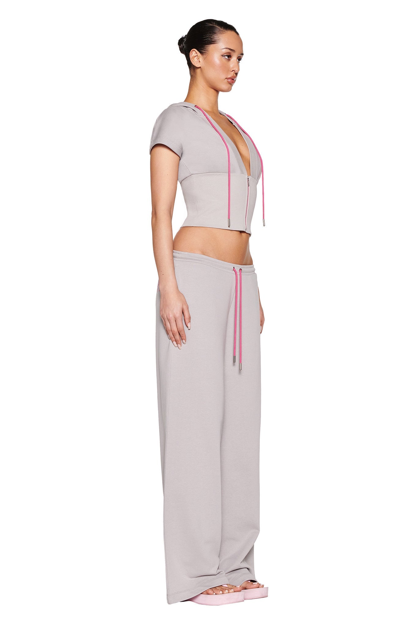 A person, shown in profile against a plain white background, wears I.AM.GIA's ARLO TRACK PANT in grey with pink accents and a light gray cropped hoodie featuring pink drawstrings. The casually chic outfit is completed with pink sandals, and their hair is styled in a bun.