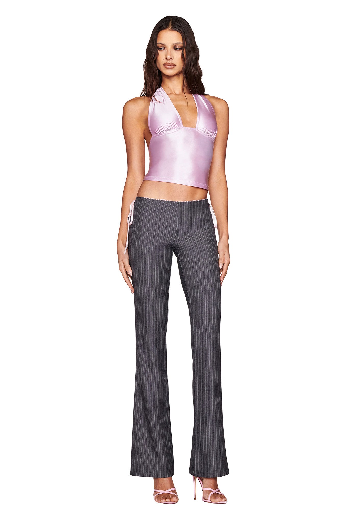 A woman confidently stands against a white background in an AURELIA HALTER TOP - PINK from I.AM.GIA, paired with pinstriped pants. Her look is completed with pink high-heeled sandals, and her long, wavy hair cascades gracefully down her shoulders.