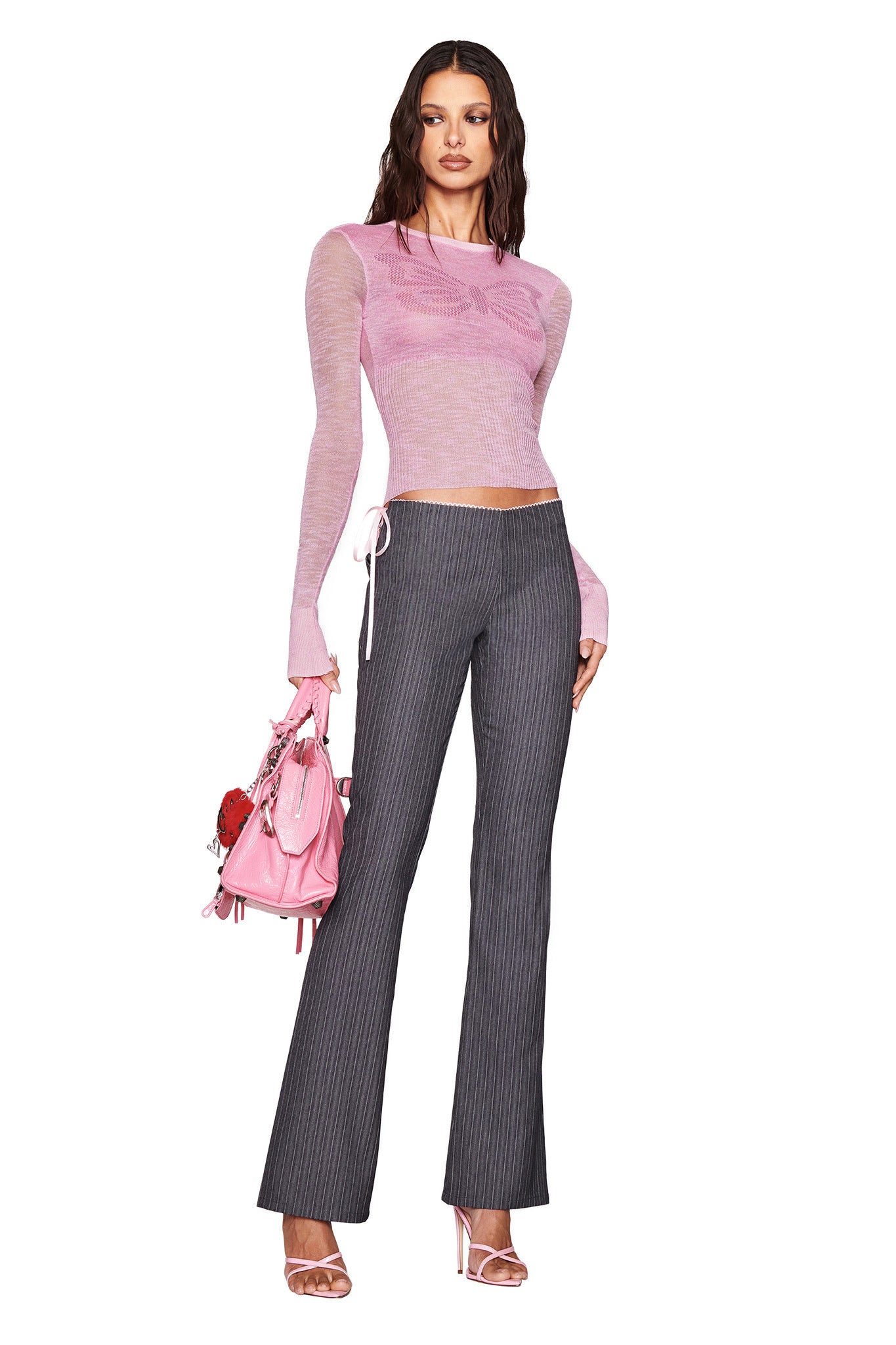 A person wearing the MARIPOSA TOP - PINK by I.AM.GIA pairs it with pinstripe gray pants, pink heels, and a pink handbag. The chic, modern outfit merges casual and formal elements against a plain white backdrop.