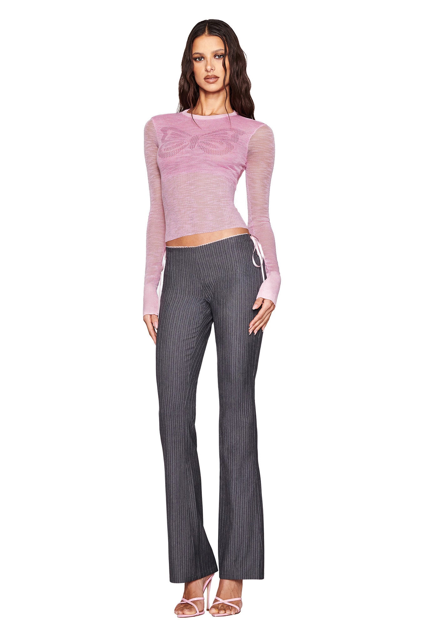 A person is standing against a white background wearing a sheer, long-sleeved pink top and the I.AM.GIA FLORIAN PANT in grey pinstripe with a tailored fit. They have wavy hair and are wearing open-toe high heels.