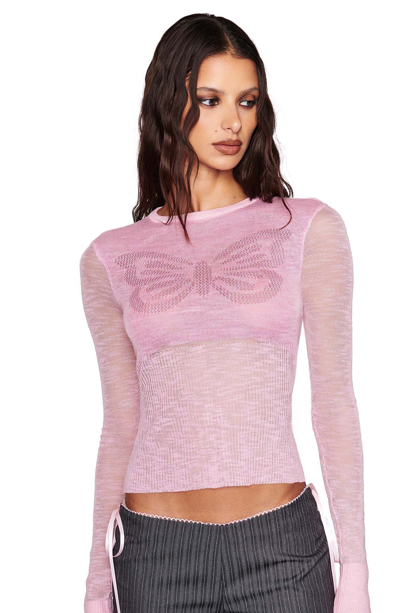 Wearing I.AM.GIA's MARIPOSA TOP in pink, featuring a sheer design and exclusive butterfly print, they pair it with gray pinstriped pants. Their long, wavy brown hair cascades as they gaze to the side against a white background.