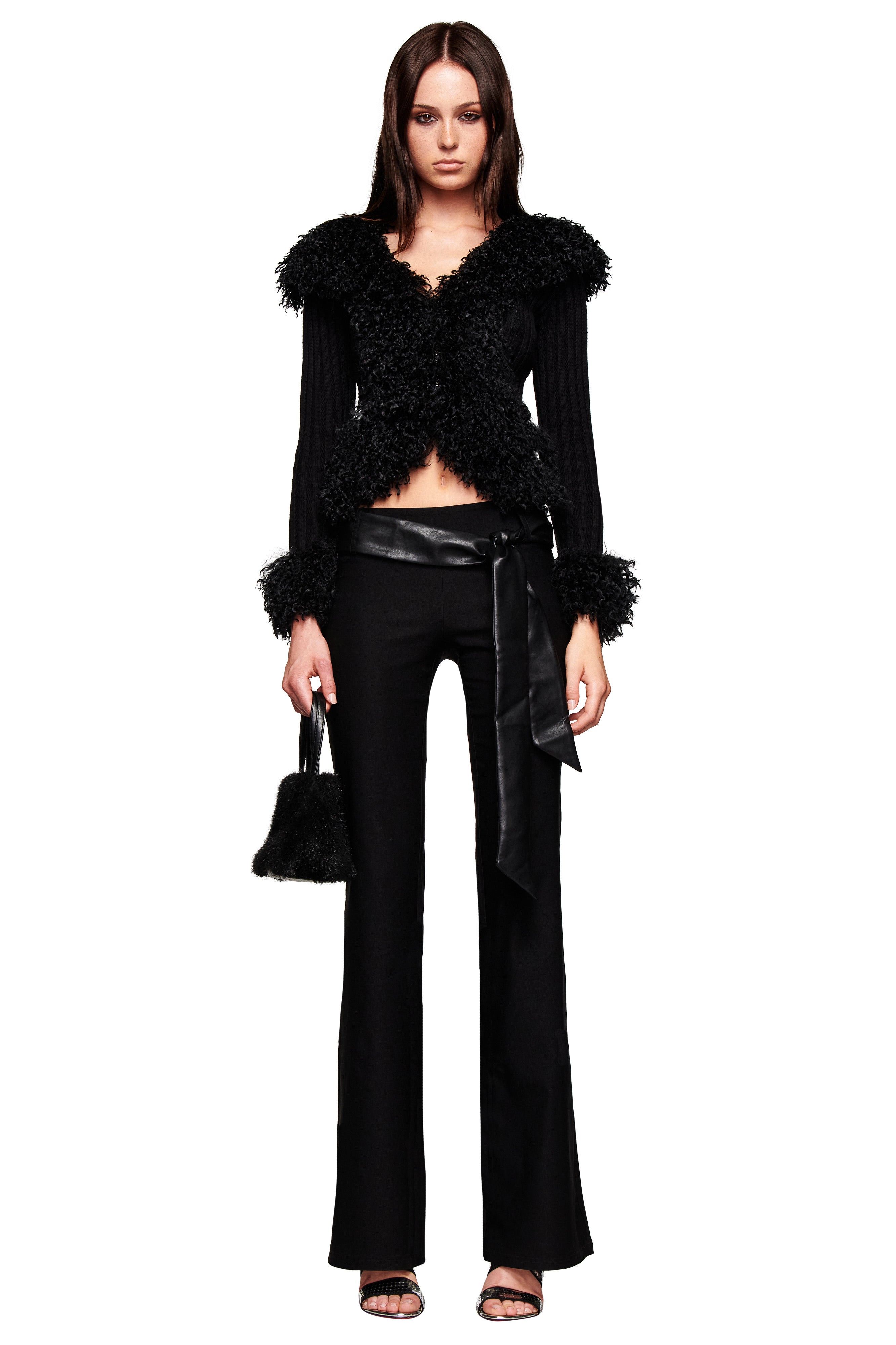 A woman faces forward wearing the I.AM.GIA FELIZ PANT in black with a textured black jacket, furry cuffs and collar, satin faux leather belt, open-toe heels, and holding a small black handbag.
