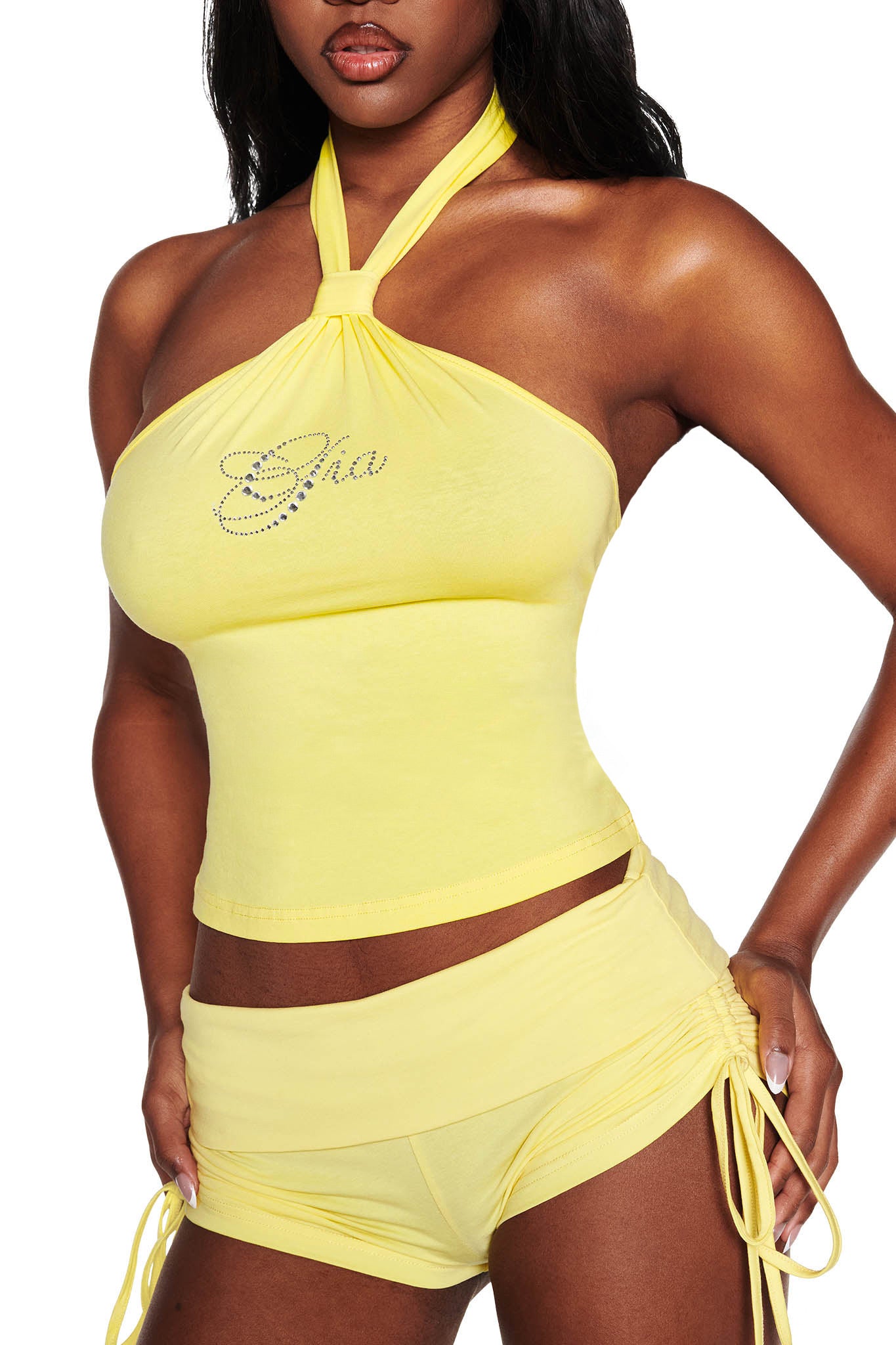Someone is wearing the ROWAN TOP - YELLOW by I.AM.GIA and matching shorts with side ties. The bright baby yellow halter top has rhinestone lettering for added sparkle, and the white background highlights the vivid ensemble.