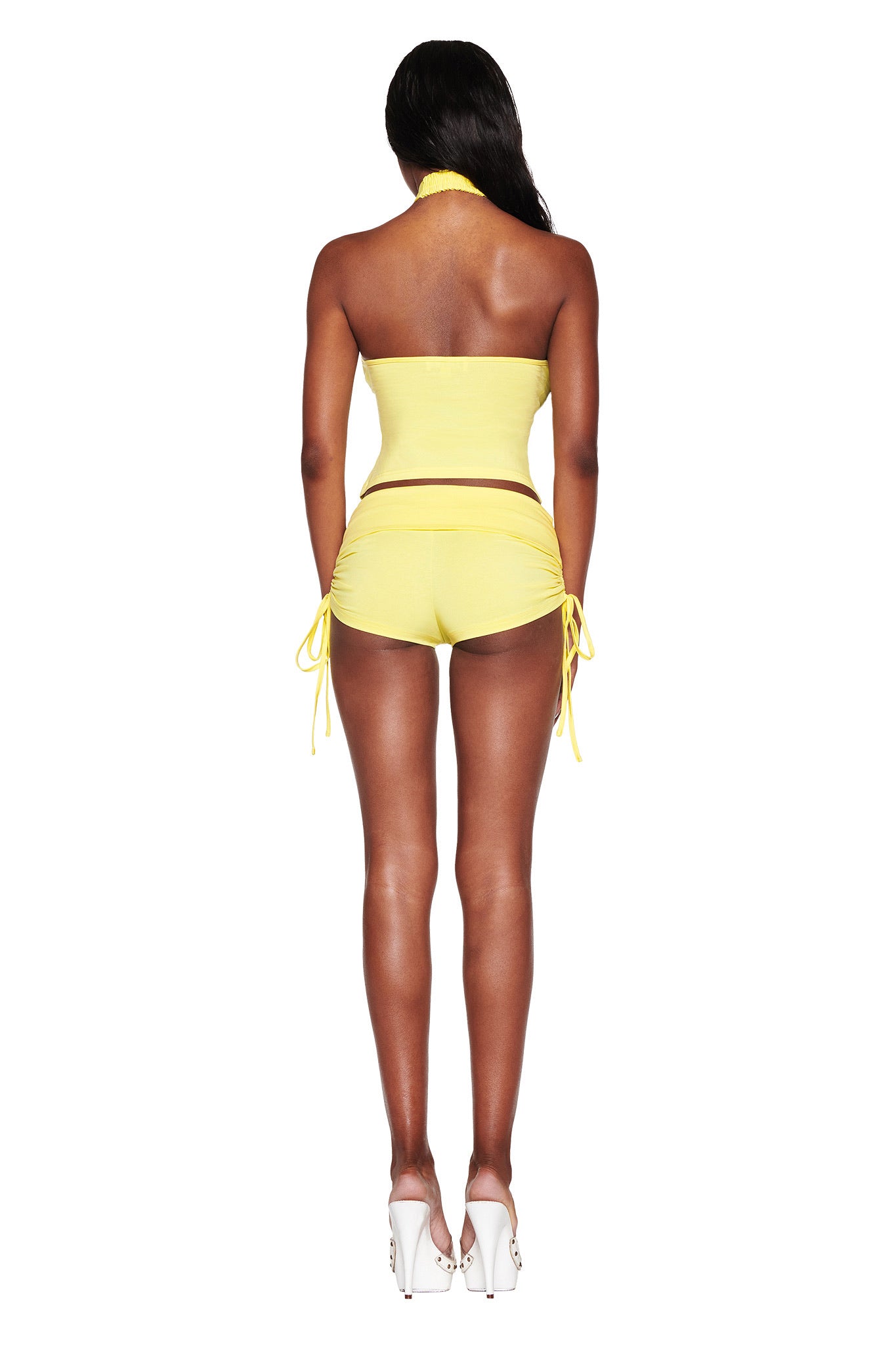 A person is seen from behind wearing the MIA SHORT in bamboo fabric by I.AM.GIA. The outfit includes a strapless top and low-rise yellow mini shorts with side ties, complemented by white high heels on a plain white background.
