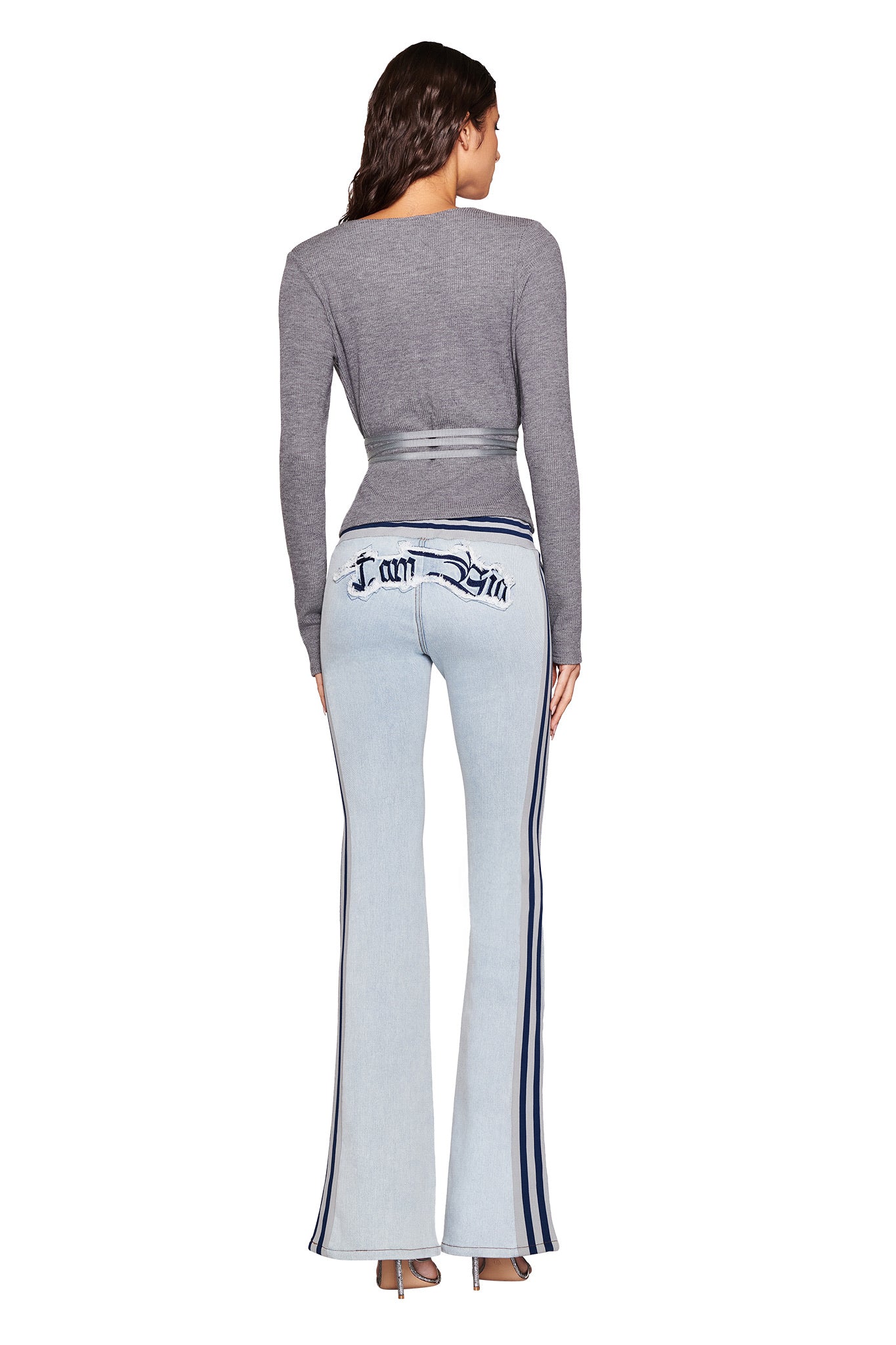 A person with long hair wears a gray long-sleeve top and I.AM.GIA's LENNOX JEAN - DENIM BLUE, featuring stylish text embroidered on the back pockets. They stand with their back to the camera, exuding an effortlessly chic vibe in these light wash denim flared pants.