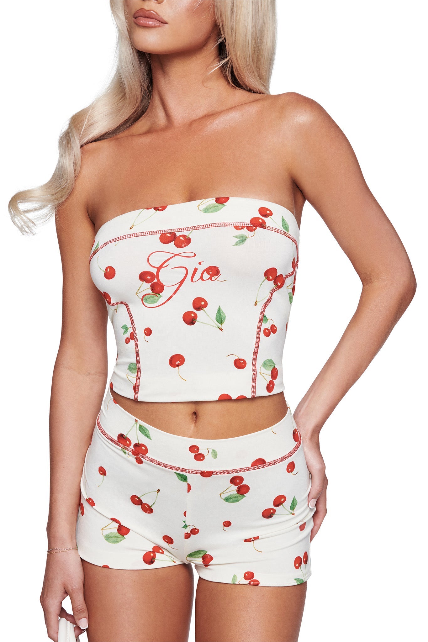 A woman wears the I.AM.GIA BLARE BOOB TUBE - CHERRY ON TOP, a white tube top with a cherry print and red stitching, paired with matching shorts. The top features “Gio” written on it. Her long blonde hair is styled down as she poses with one hand by her side.
