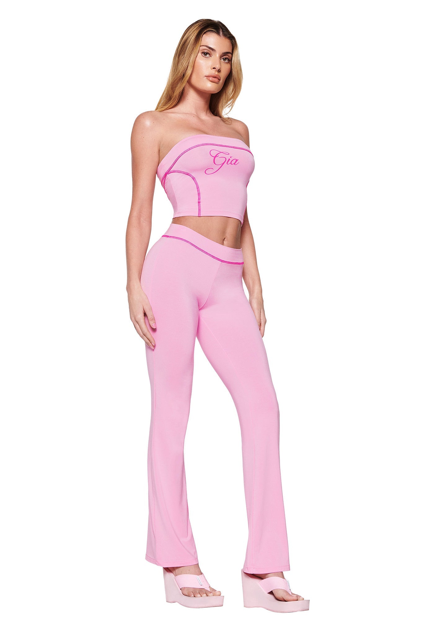 Someone wearing the BLARE BOOB TUBE - PINK in bubblegum, paired with matching high-waisted flared pants and pink platform sandals, stands against a white background. The I.AM.GIA logo is featured on the front amid curved patterns and bold contrast stitching.