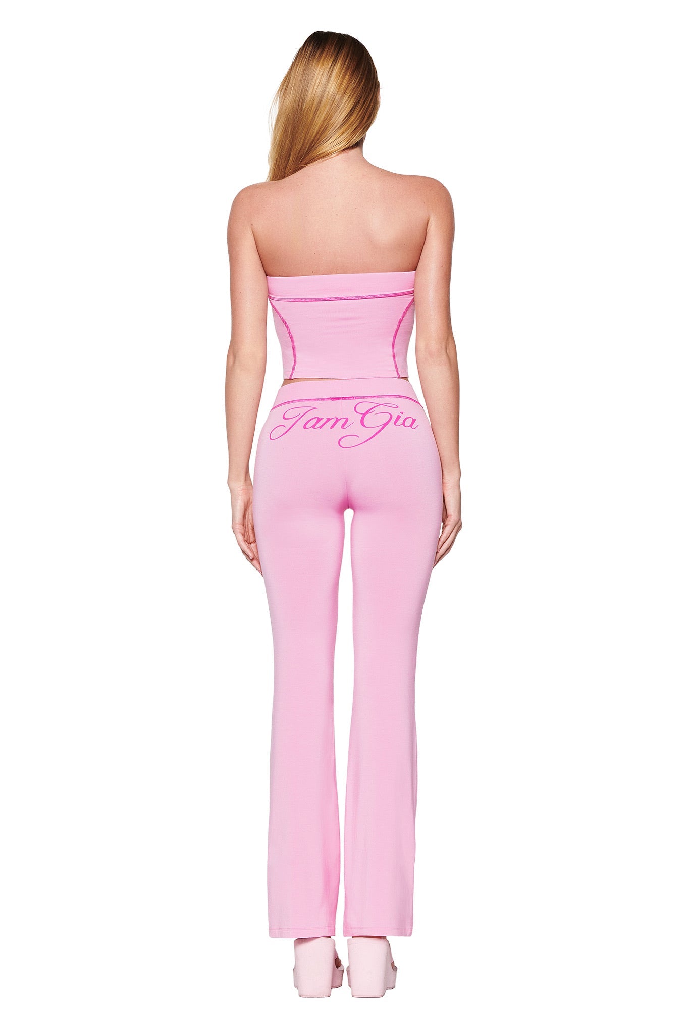A rear view of a person with long hair wearing BLARE TRACKPANT in bubblegum pink by I.AM.GIA, featuring the bold logo on the back, paired with matching pink platform shoes, confidently standing against a white background.