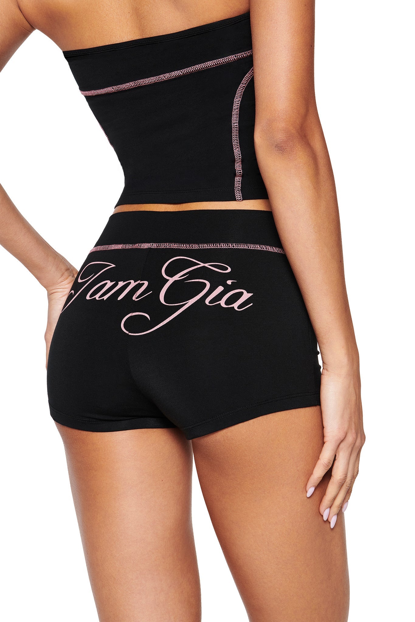The ensemble showcases a chic bodycon fit with a black strapless top and BLARE SHORT - BLACK featuring pink stitching. "I.AM.GIA" elegantly written in large pink cursive on the shorts complements the style, as the person’s left arm rests confidently on their hip.