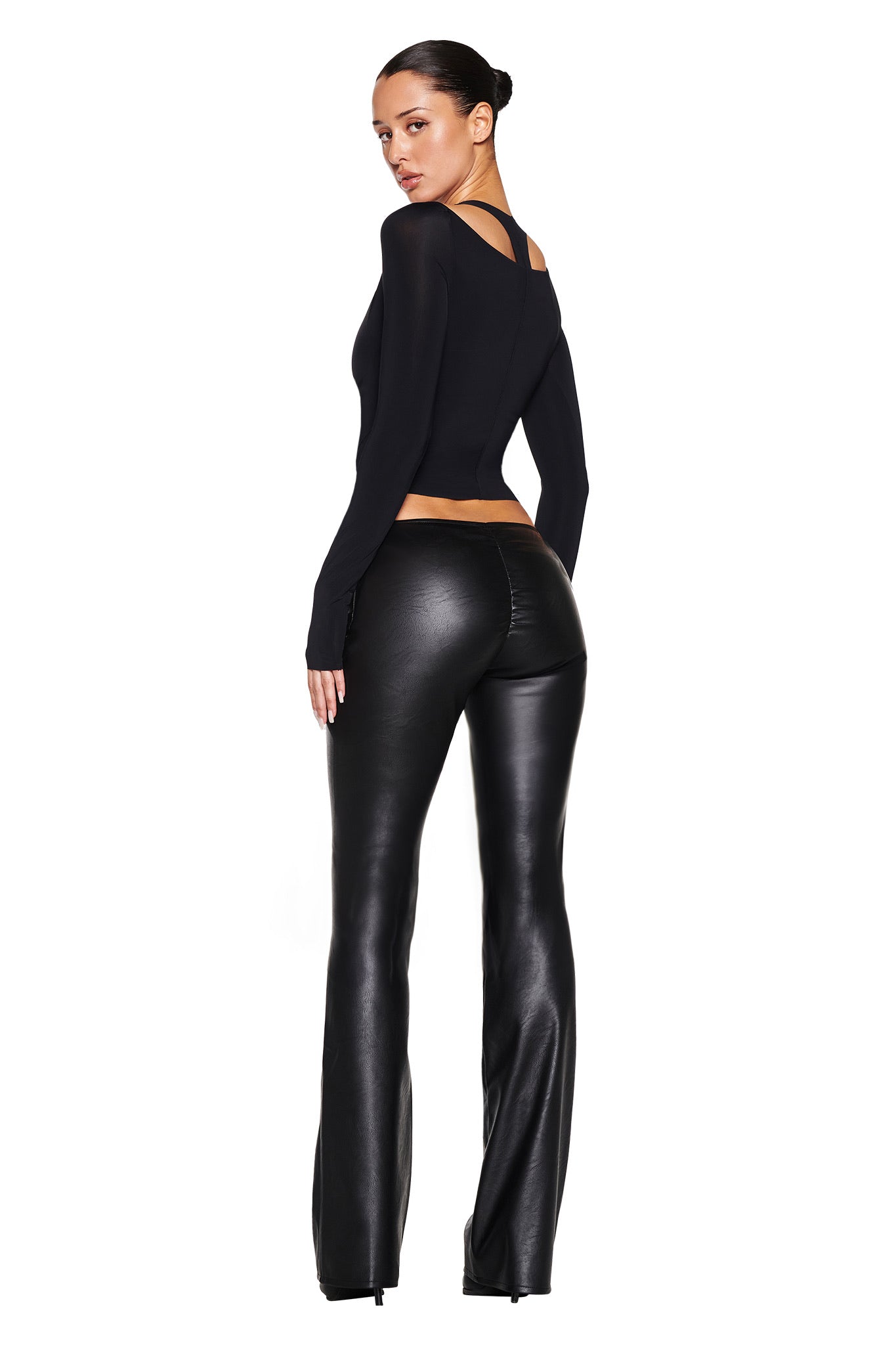 A person models high-waisted black leather pants paired with the NERINA LONG SLEEVE TOP - BLACK from I.AM.GIA, featuring an open back design. Posed against a white backdrop, the outfit's chic appeal and high stretch fabric are emphasized as they glance over their shoulder.