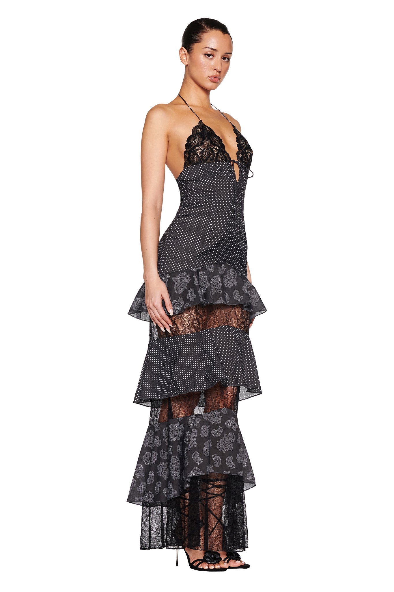 Against a white background, a woman models the I.AM.GIA PEARL MAXI DRESS in black polka dot. This long layered gown features lace, sheer patterns, a halter neckline, and tiered ruffles. She completes her look with strappy heels and an elegant updo.