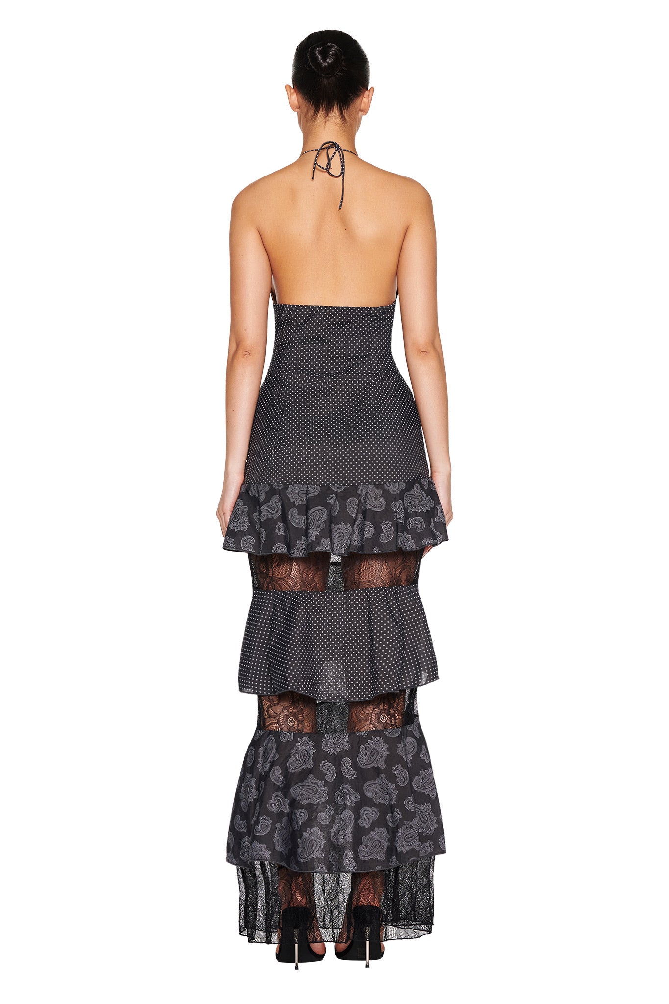 Rear view of a person in the I.AM.GIA PEARL MAXI DRESS - BLACK : POLKA DOT, featuring a fitted bodice and tiered skirt with lace panels. The backless design adds elegance, complemented by black heels.