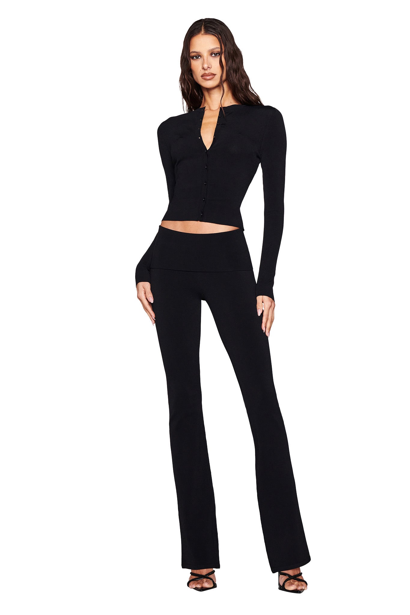 Wearing the I.AM.GIA Audrina Cardigan 2.0 in black, along with flared pants, she exudes confidence. Her long wavy hair cascades as she poses in high-heeled sandals against a plain white background.