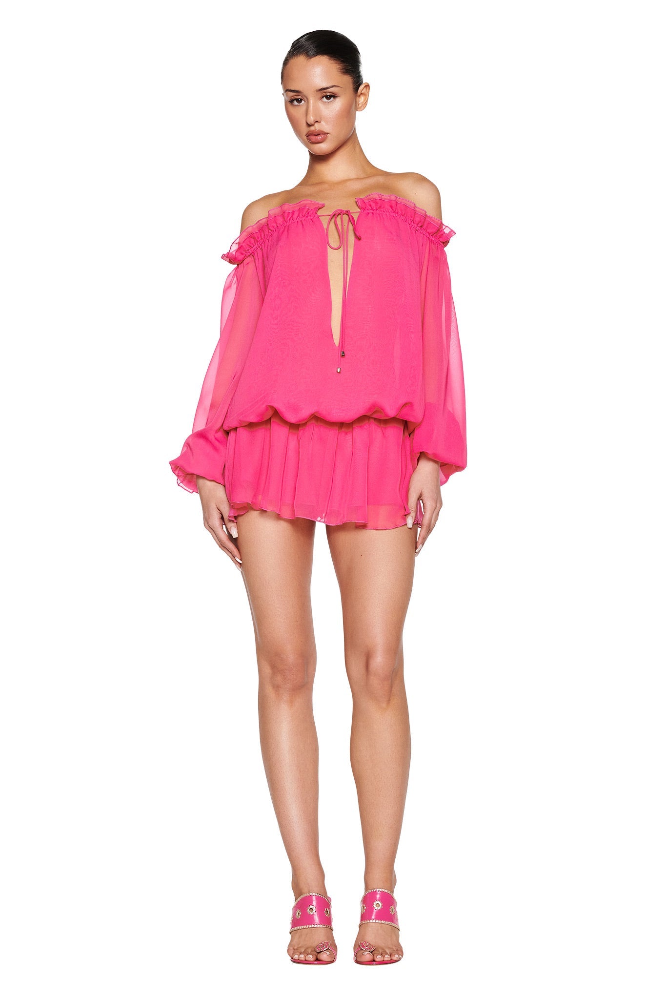 Someone poses in the JAZMIN PLAYSUIT - PINK by I.AM.GIA, featuring a hot pink georgette, off-the-shoulder design with long sleeves and a ruffled neckline, paired with matching pink heels against a plain white backdrop.