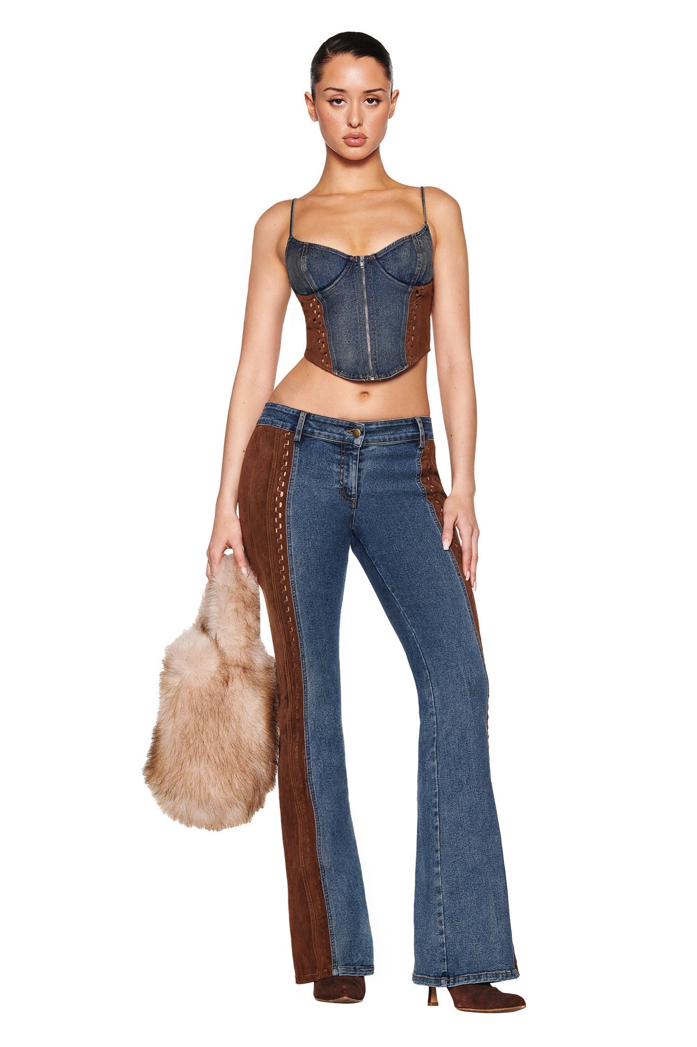 A woman models I.AM.GIA's FORREST BUSTIER in medium wash denim, paired with flare jeans featuring brown suede panels. She holds a tan faux fur bag, wears brown shoes, and has her hair tied back against a plain white background.