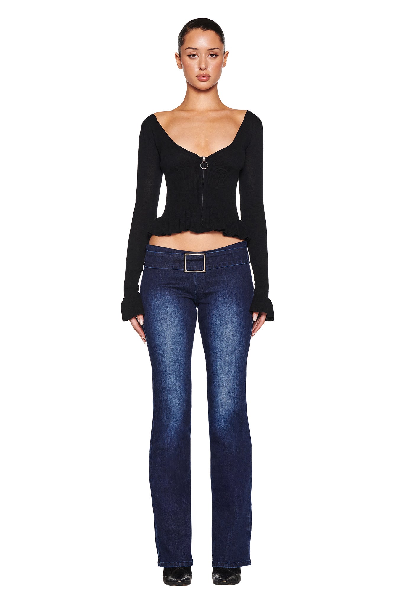 Facing forward against a white backdrop, the individual wears the RAVEN TOP in black by I.AM.GIA with dark blue jeans secured by a belt. They have black shoes and upward-styled hair.