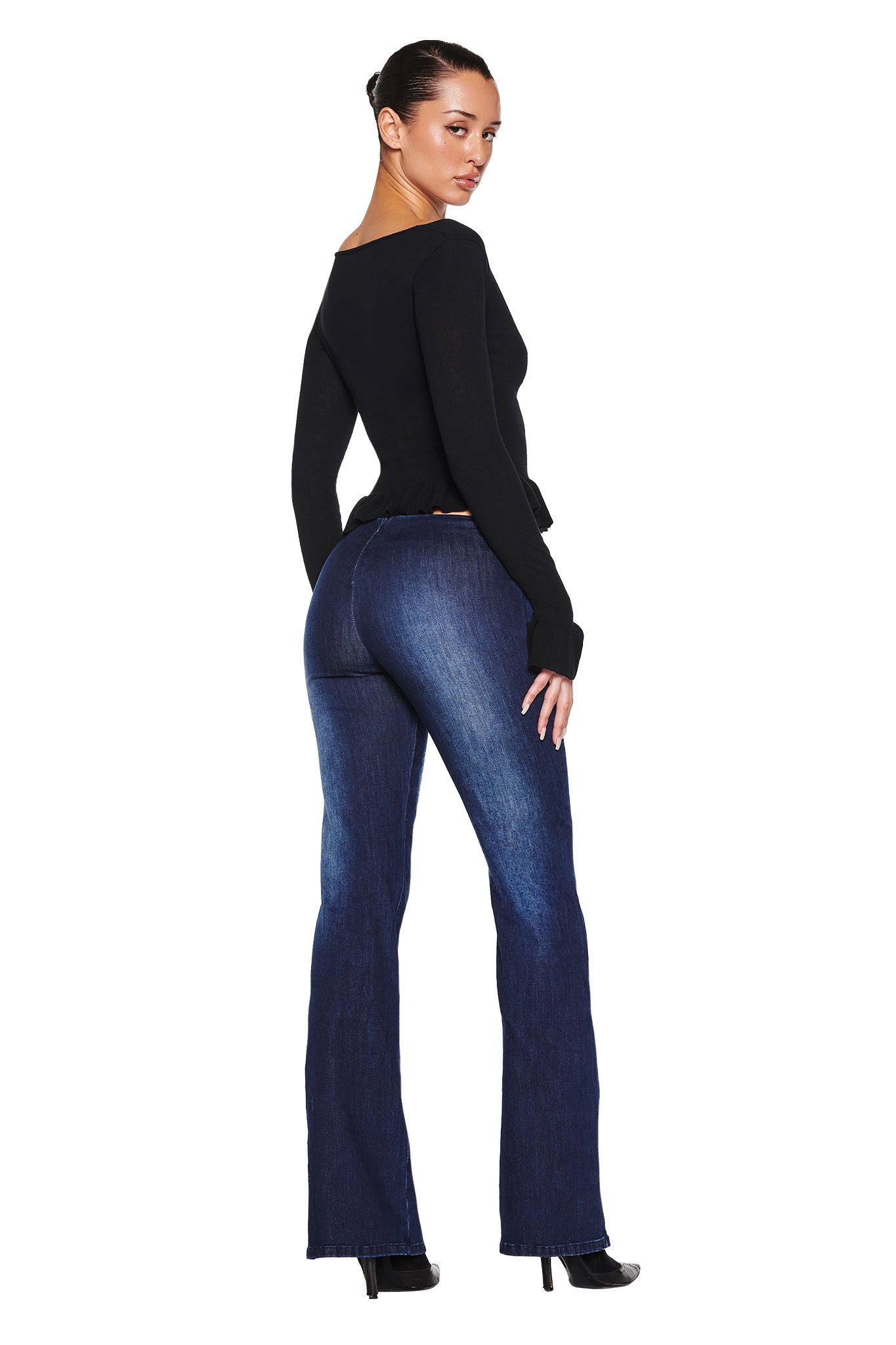 A person confidently stands against a white background, wearing the RAVEN TOP - BLACK by I.AM.GIA, with dark hair pulled back, styled alongside dark blue flared jeans and black heels. Their back faces the camera.