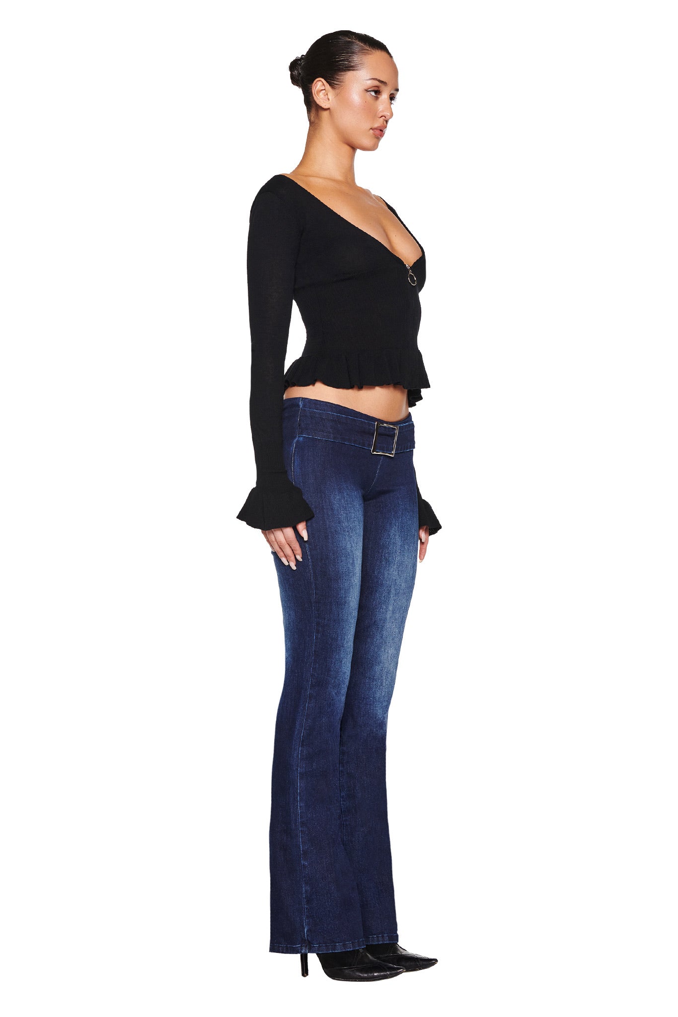 Side view of a person wearing the I.AM.GIA RAVEN TOP - BLACK with frilled cuffs and dark blue bootcut jeans, standing against a white background. Their hair is tied back, hands at their sides, exuding effortless style synonymous with timeless knit fashion.