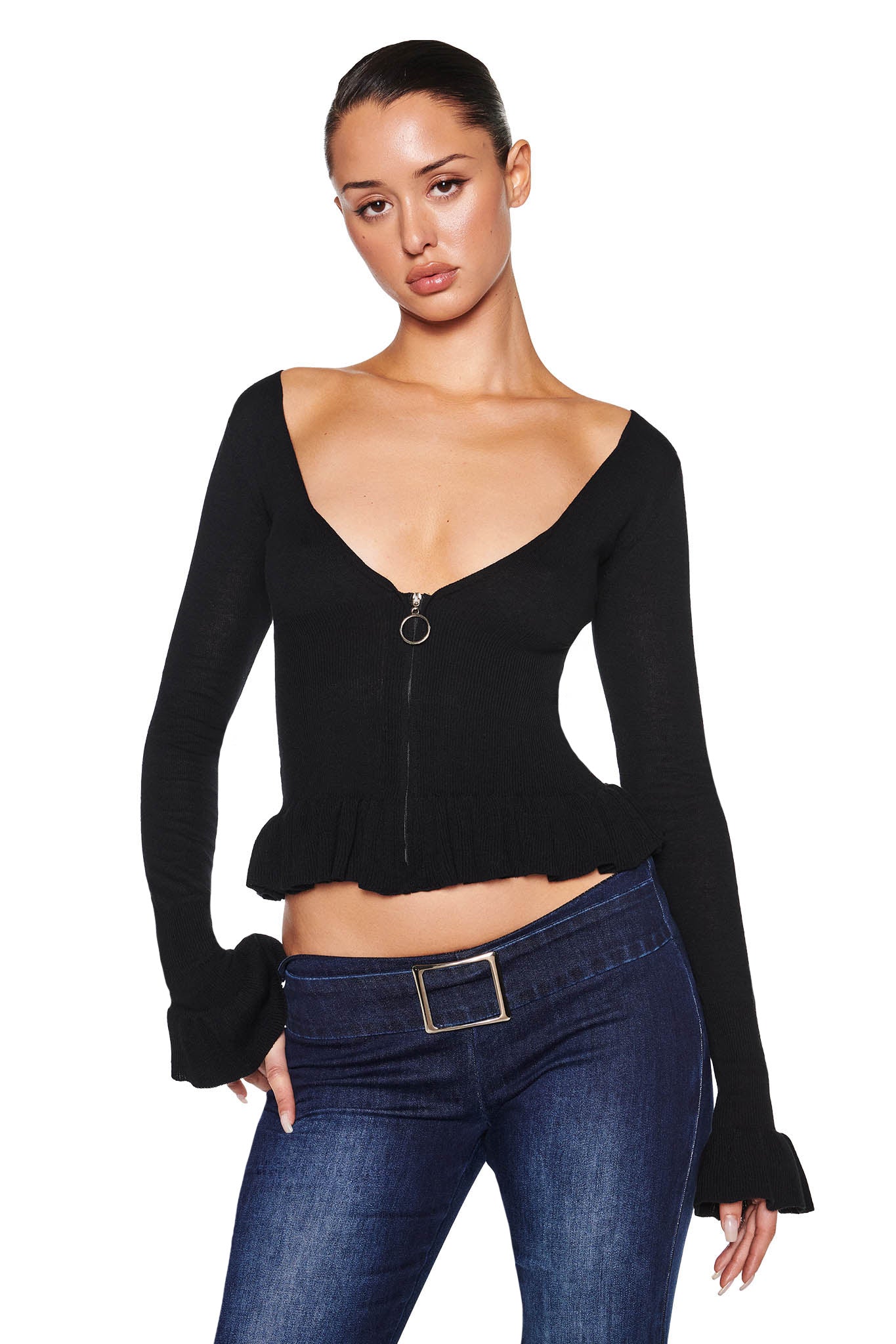 A woman wears the I.AM.GIA RAVEN TOP in black, a fitted long-sleeve knit sweater featuring a deep V-neckline and silver zipper, with a ruffled hem. It pairs perfectly with dark blue jeans cinched by a belt with a rectangular buckle against a plain white backdrop.