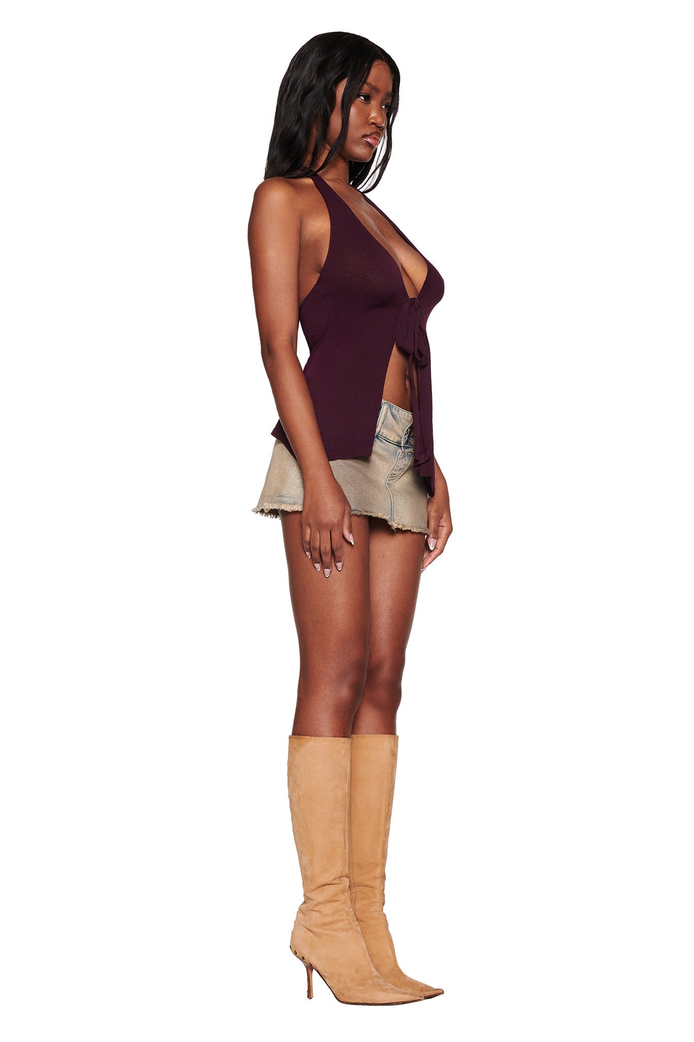 A woman in a SIMI TOP - PURPLE : PLUM by I.AM.GIA and beige shorts stands in profile with tan knee-high heeled boots. Her dark, wavy hair cascades gracefully as she poses against a plain white background.
