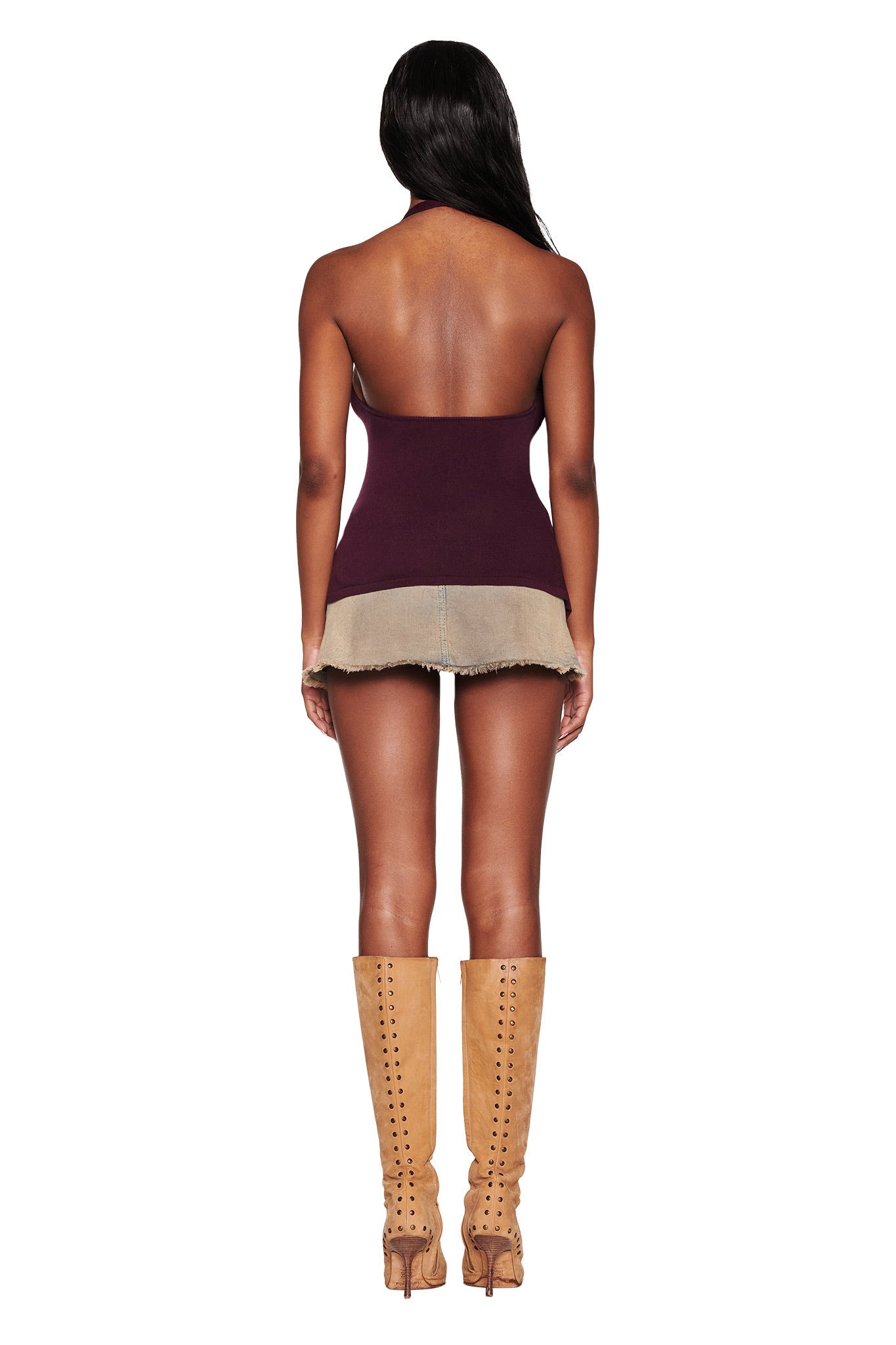 A woman stands against a white background, showcasing the I.AM.GIA SIMI TOP in plum purple, featuring a plunging V neckline. She pairs it with a short beige skirt and knee-high tan heeled boots.