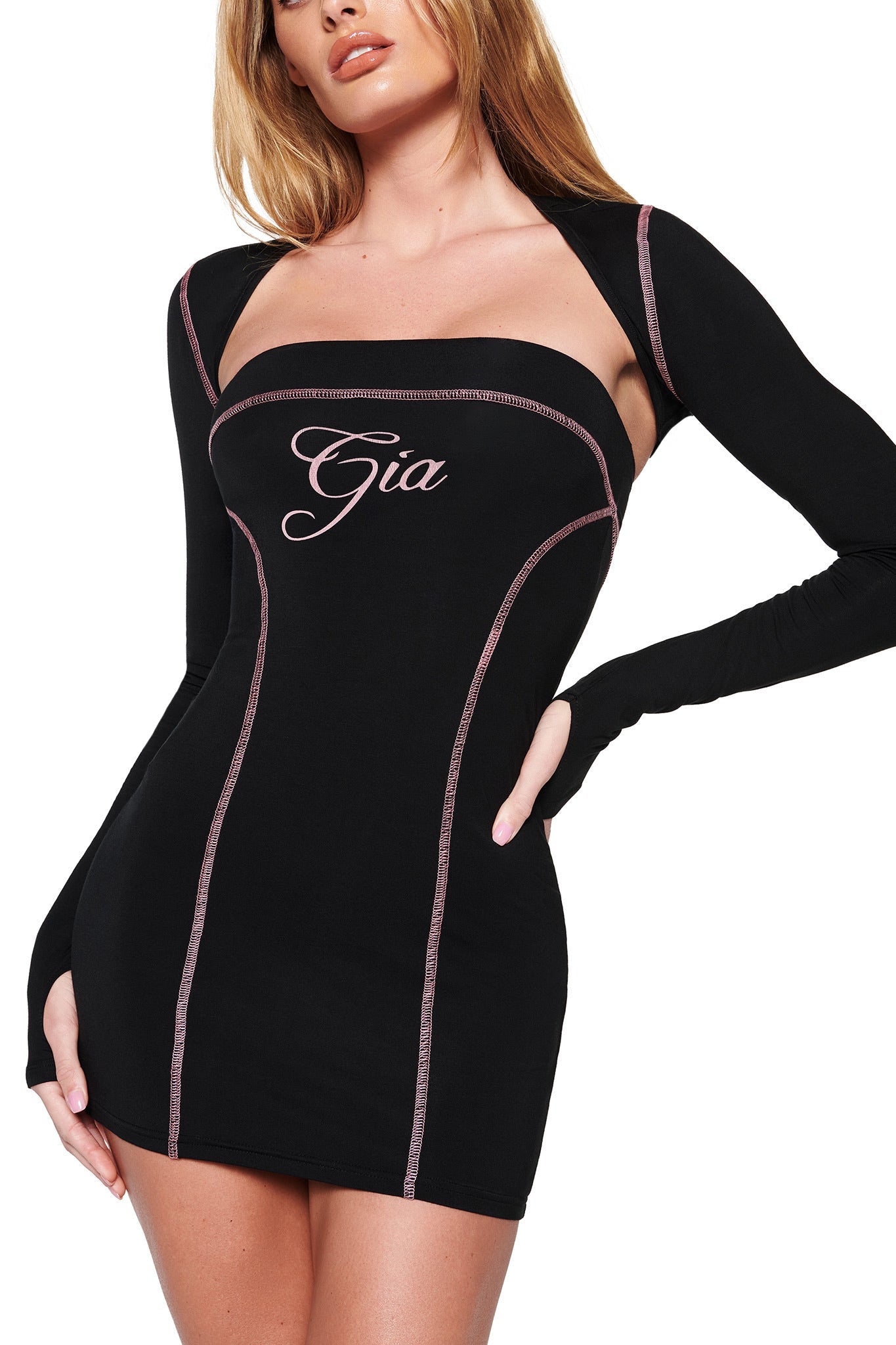 A woman elegantly poses against a white backdrop, showcasing the I.AM.GIA BLARE SHRUG in black. This form-fitting mini dress features distinctive contrast stitching and pink embroidery spelling "Gia," accentuated by pink stitching along the seams.
