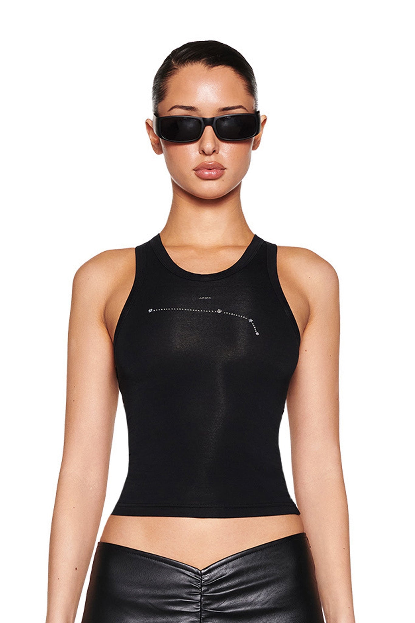 A person stands against a white background, styled in I.AM.GIA's ARIES ZODIAC TANK - BLACK paired with leather pants. With dark sunglasses and slicked-back hair, they achieve a sleek, modern look.
