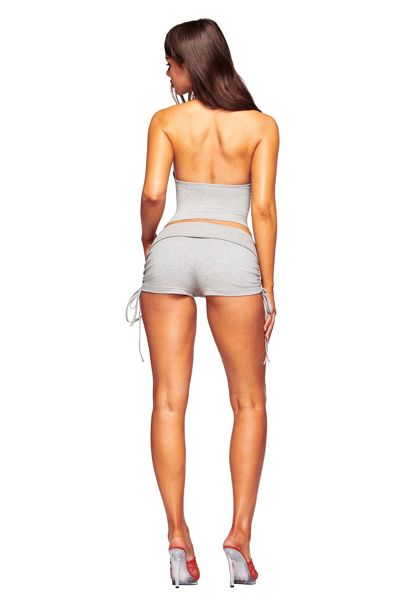 A woman with long brown hair, facing away, wears the I.AM.GIA ROWAN TOP in heather grey as a strapless tube top with gray drawstring shorts and clear heels, set against a white background.