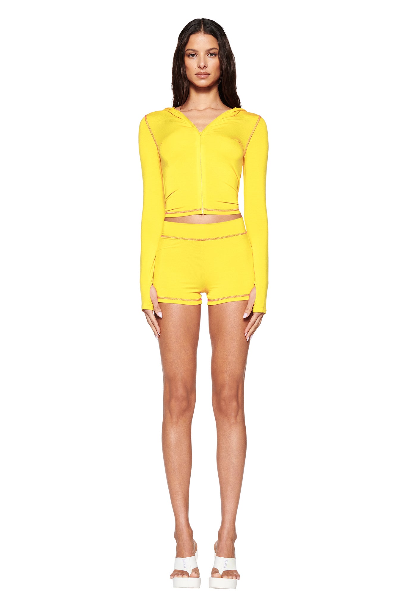 Against a white background, a person showcases the I.AM.GIA ensemble, including the vibrant lemon BLARE SHORT with a bodycon fit and matching zip-up hoodie. They pair it with white platform sandals, while their long hair flows freely, enhancing the chic vibe.