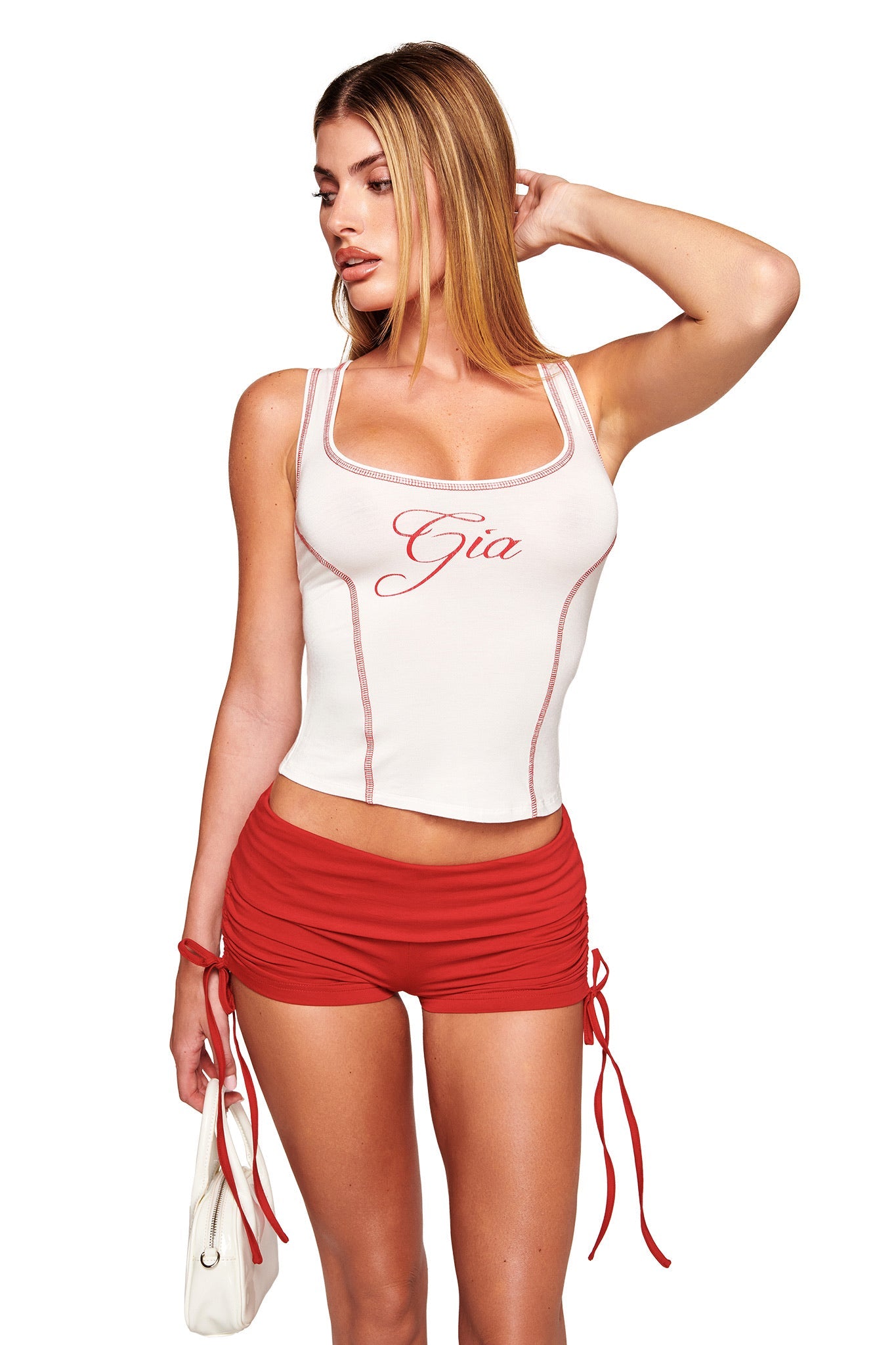 A woman with long blonde hair wears a fitted white "Gia" script tank top and I.AM.GIA MIA SHORT - RICH RED ruched low rise mini shorts, holding a small white purse, posing with her right hand in her hair against a white background.