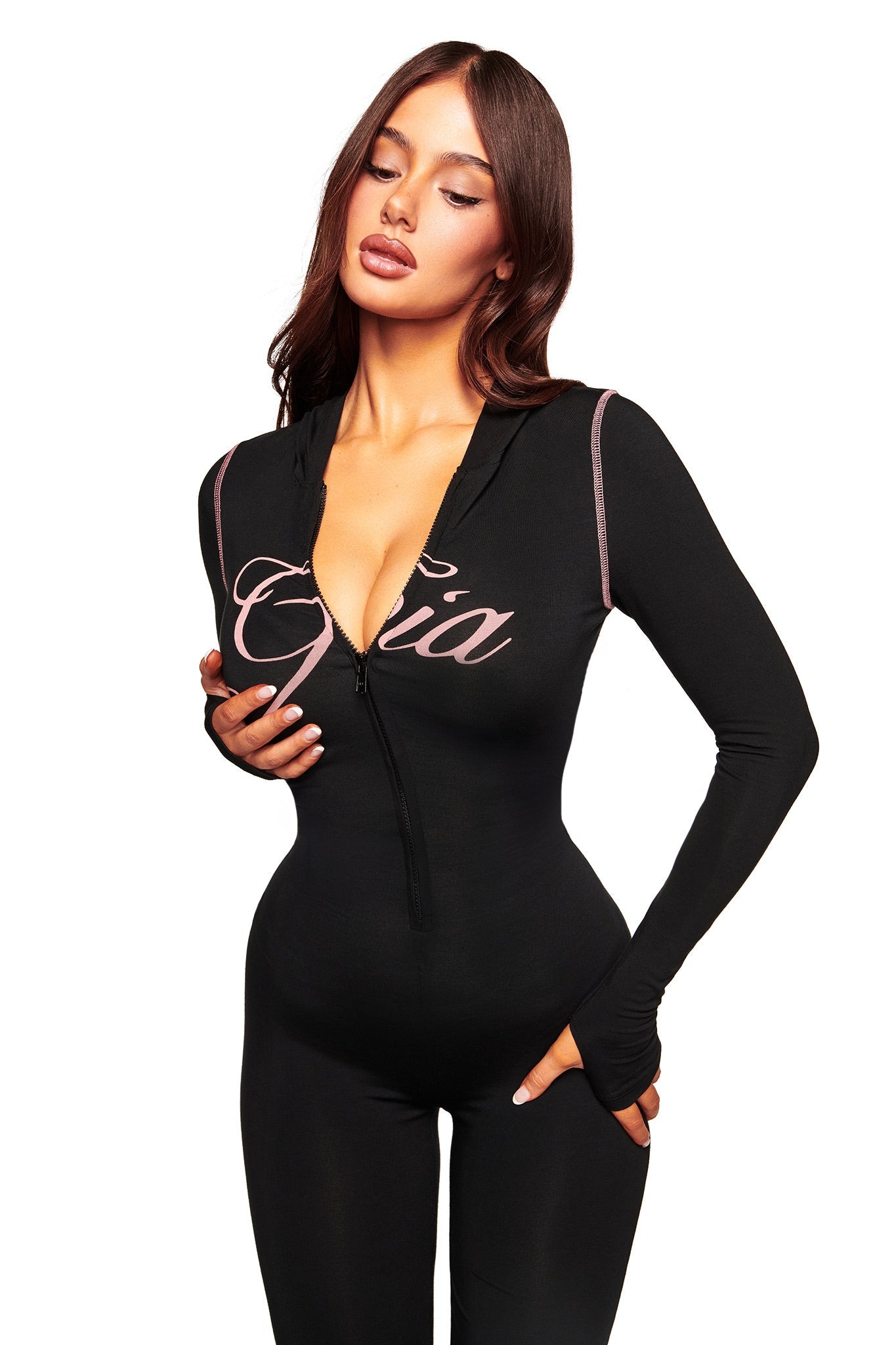 A woman in the I.AM.GIA BLARE JUMPSUIT—black, fitted, long-sleeve, hooded with a front zipper, flared leg, and "Cía" script on the chest—stands against a white background.