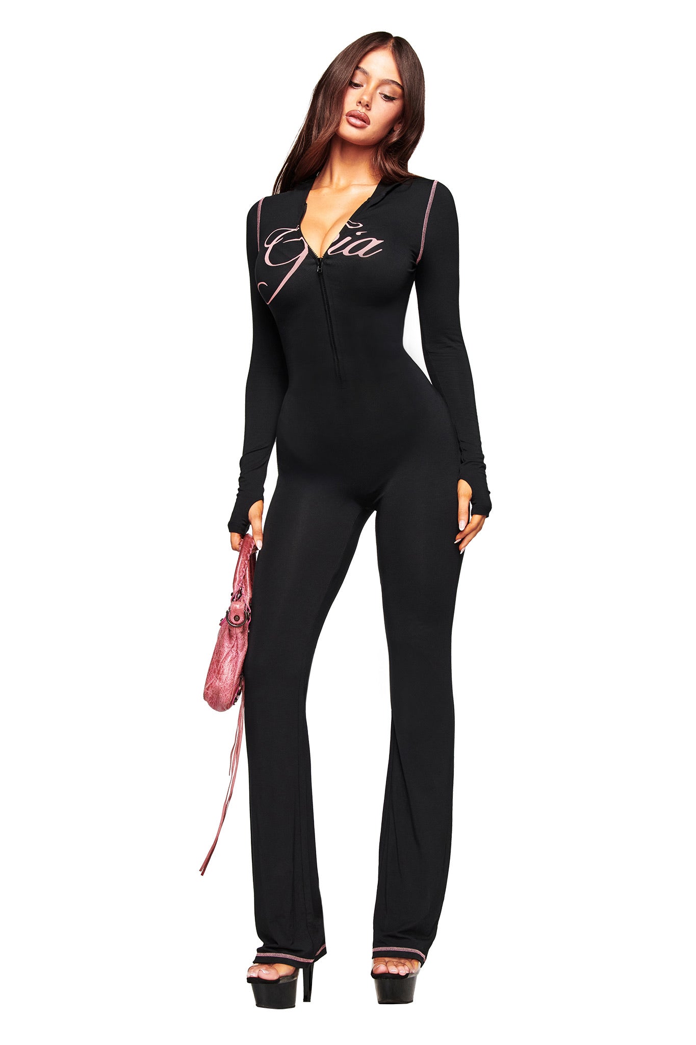 A woman with long brown hair showcases the I.AM.GIA BLARE JUMPSUIT - BLACK, featuring pink trim and “GIA” on the chest. She pairs it with high platform heels and a pink handbag, highlighting its flared leg against a white background.