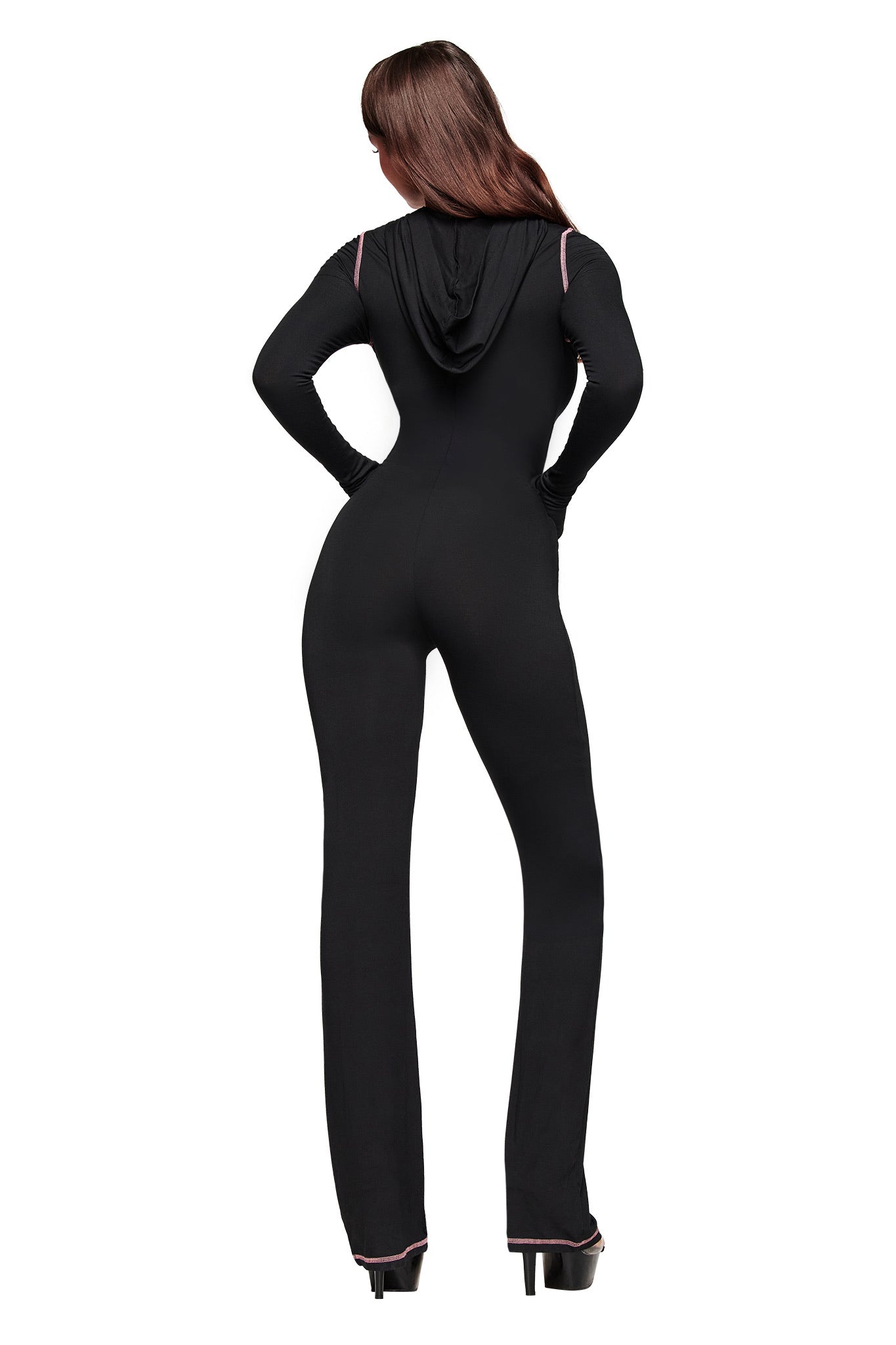 A person with long brown hair, wearing the I.AM.GIA BLARE JUMPSUIT - BLACK with flared legs and a hood, stands facing away against a white background, hands on hips.