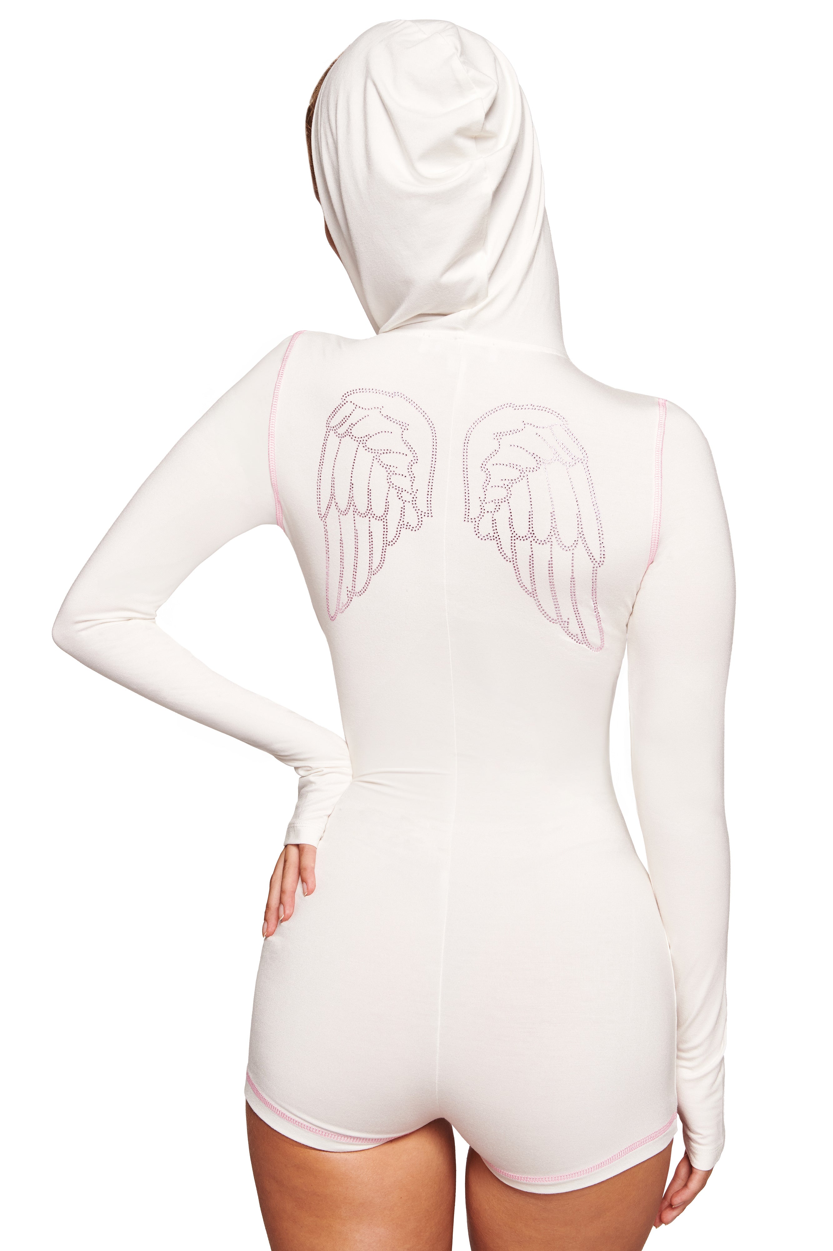 A person wears the I.AM.GIA BLARE ROMPER - WHITE ANGEL, a mini white bodycon romper with long sleeves, hood, light pink trim, and pink-outlined angel wings on the back, shown from behind for a stylish effect.